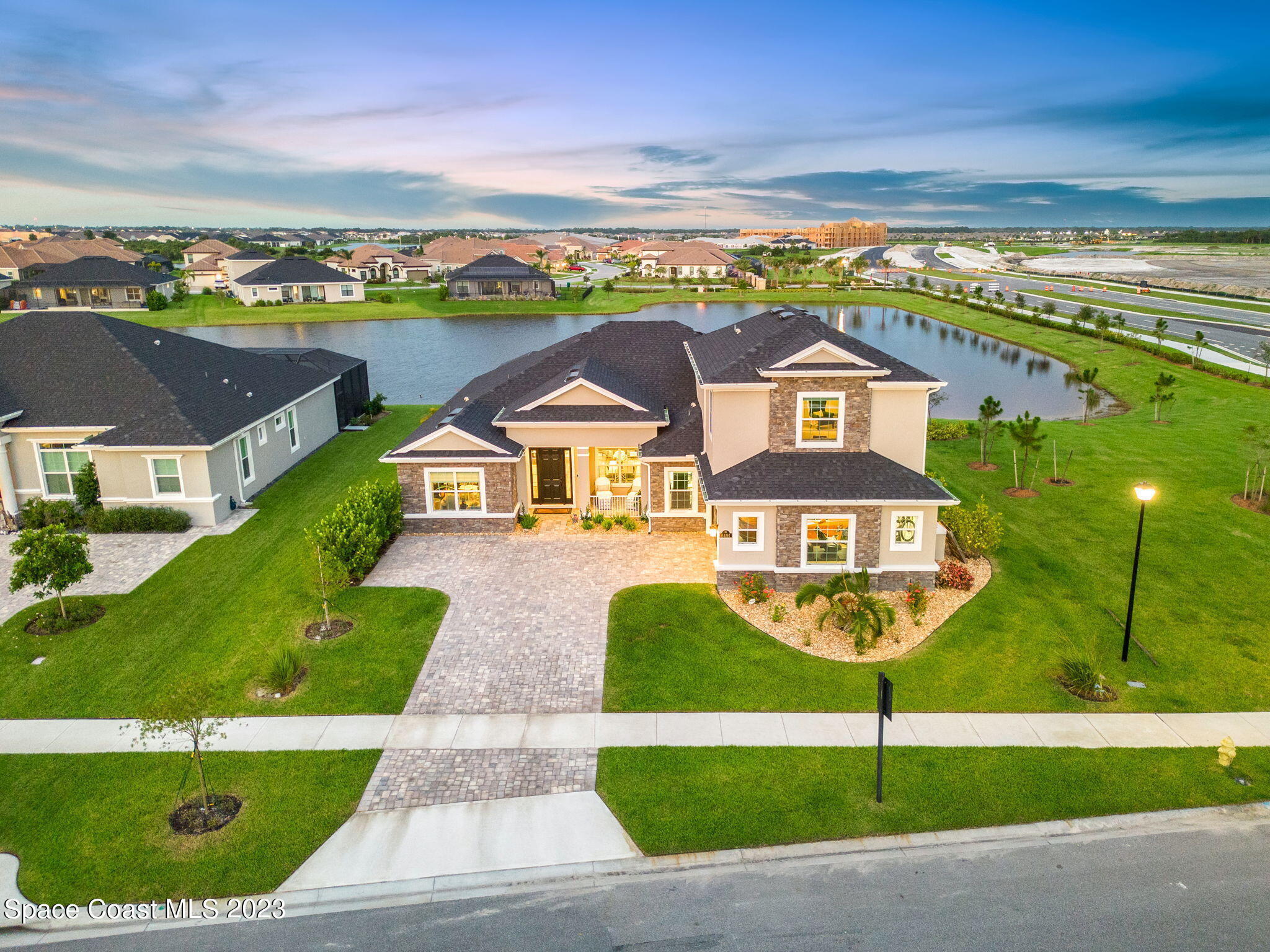 Two-story beige and stone home beside a pond, seen from above with its landscaped front lawn and driveway.