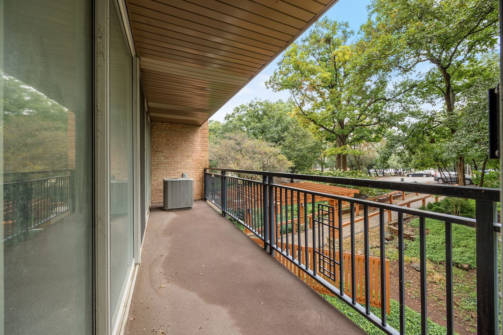 Covered condo balcony with black railing overlooking mature trees and landscaped grounds.