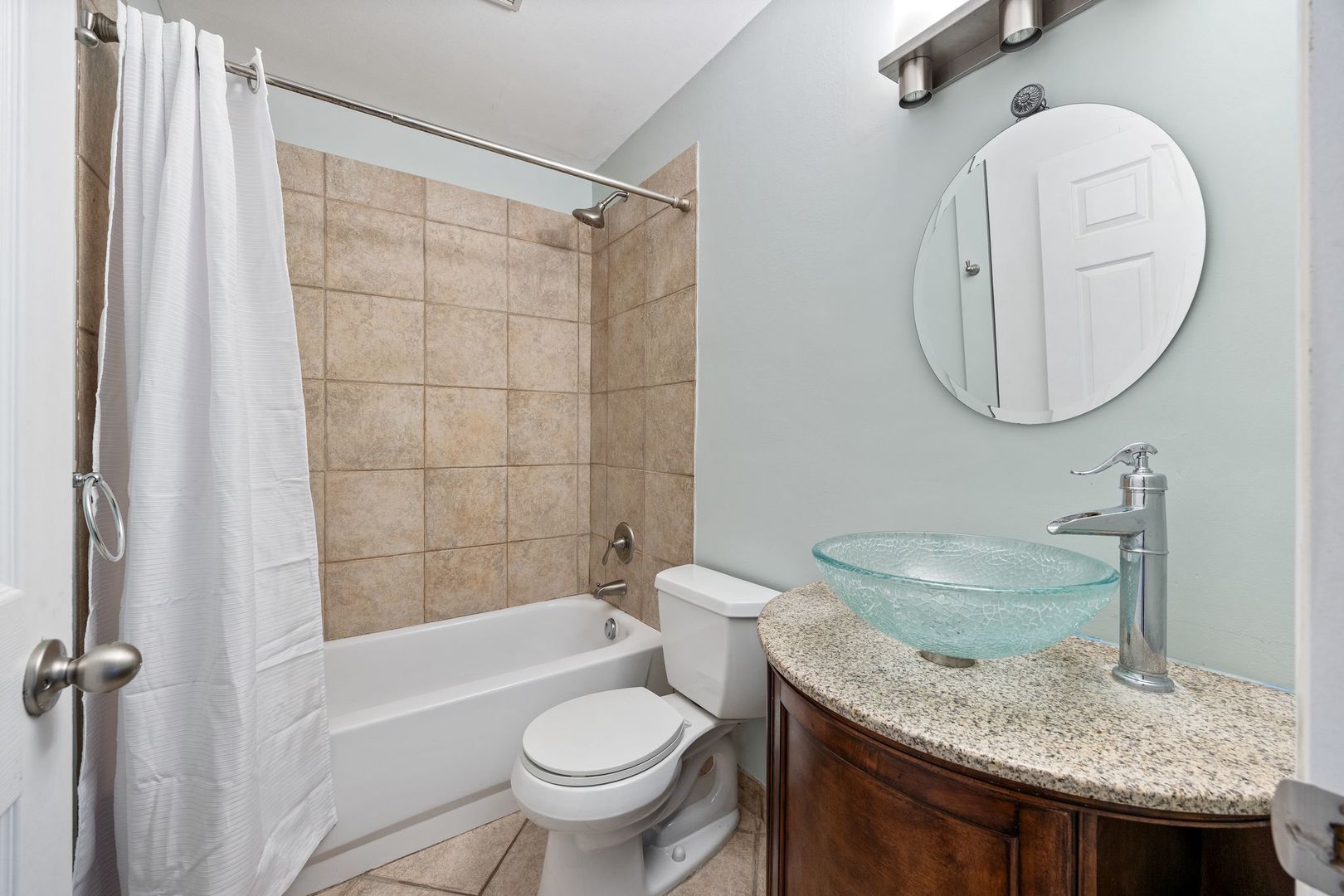 Bathroom with a turquoise glass vessel sink, granite countertop, and beige tiled tub surround.