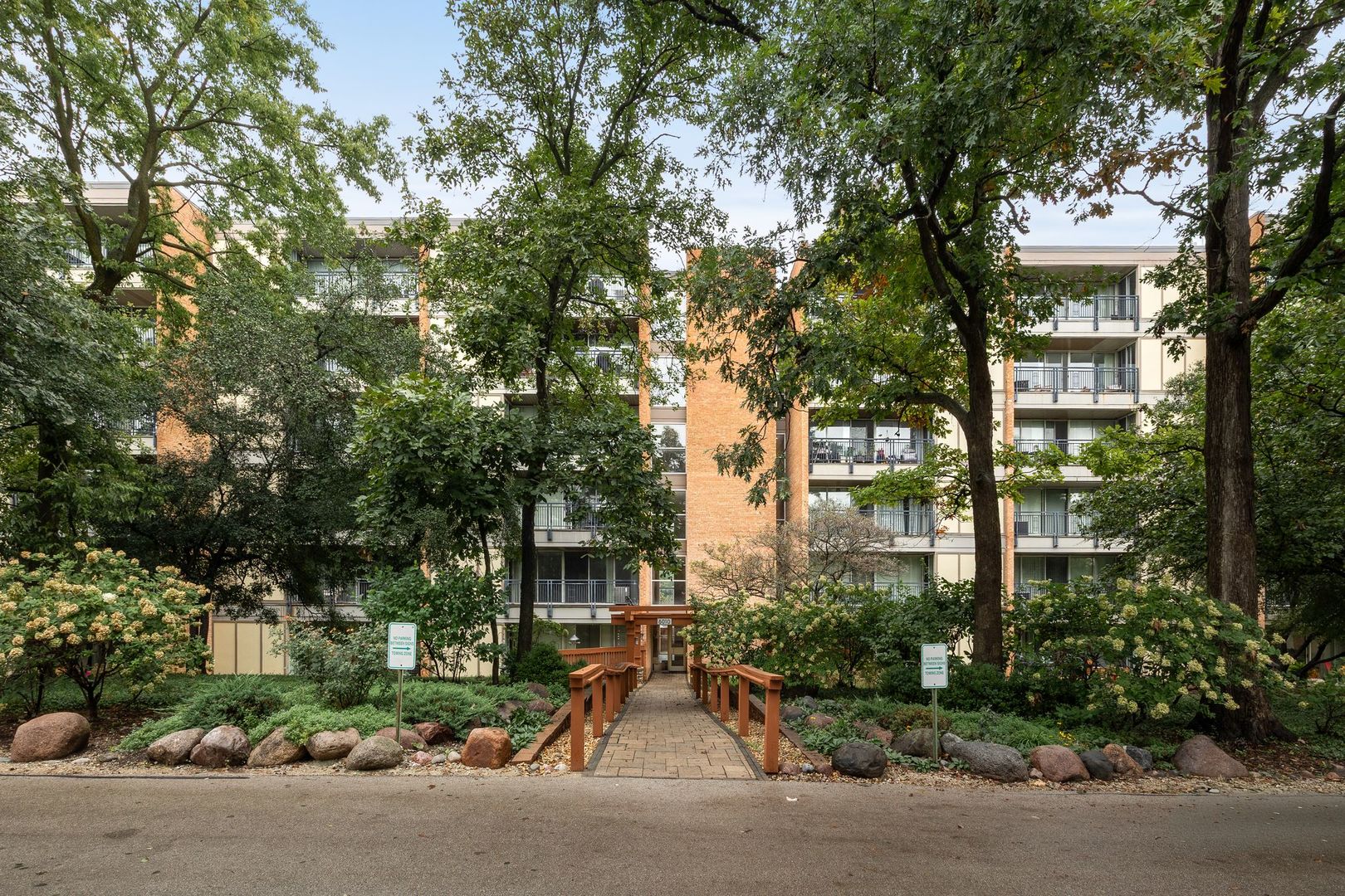 Condominium building with balconies behind a landscaped walkway and mature trees.
