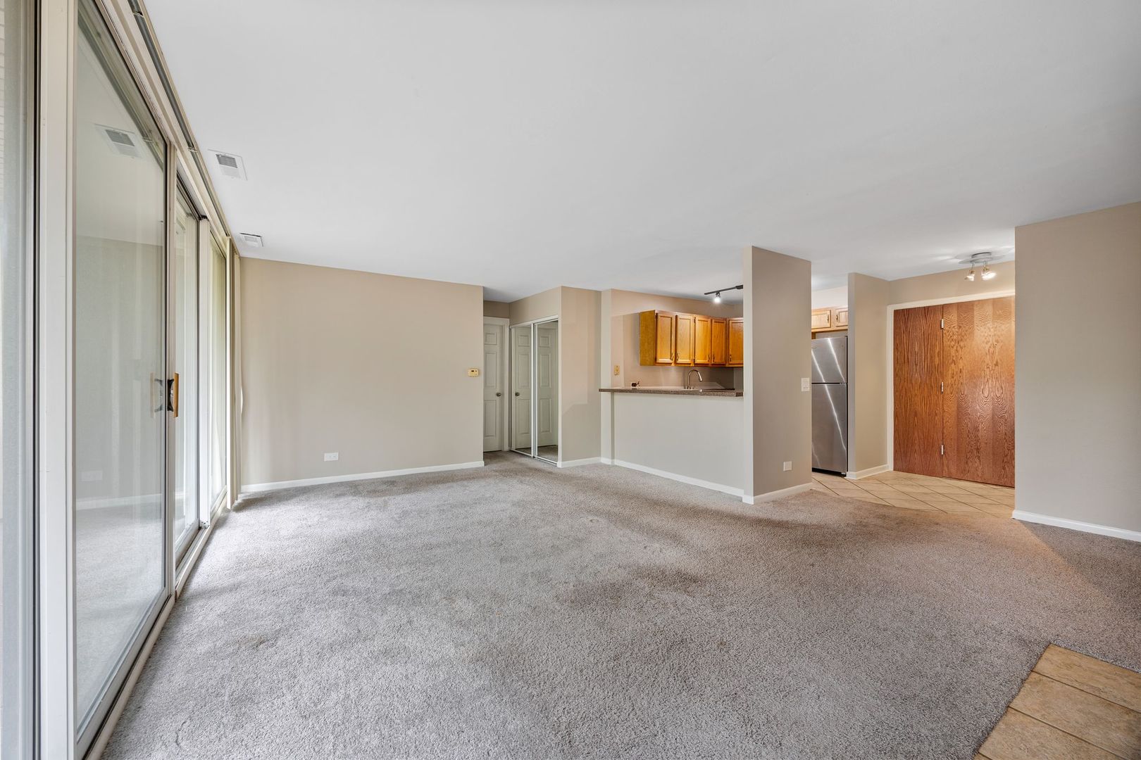 Empty living area with gray carpet, sliding glass doors, and an open kitchen with wood cabinets.