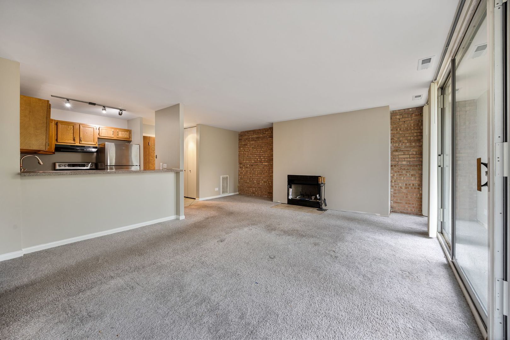 Empty condo living room with a brick fireplace, open kitchen, and large sliding glass doors.