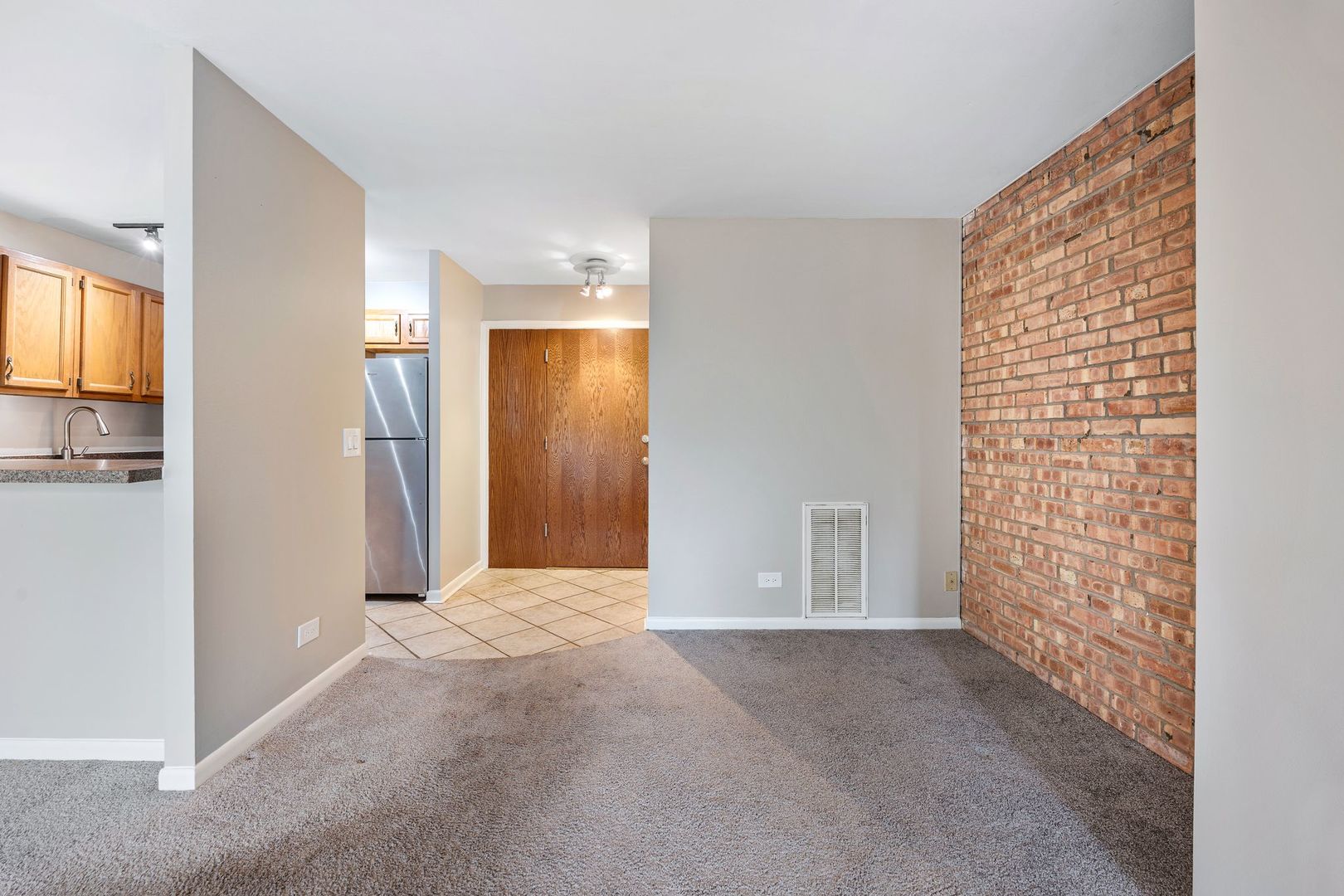 Empty living room with a brick accent wall, gray carpet, and a view toward the kitchen.