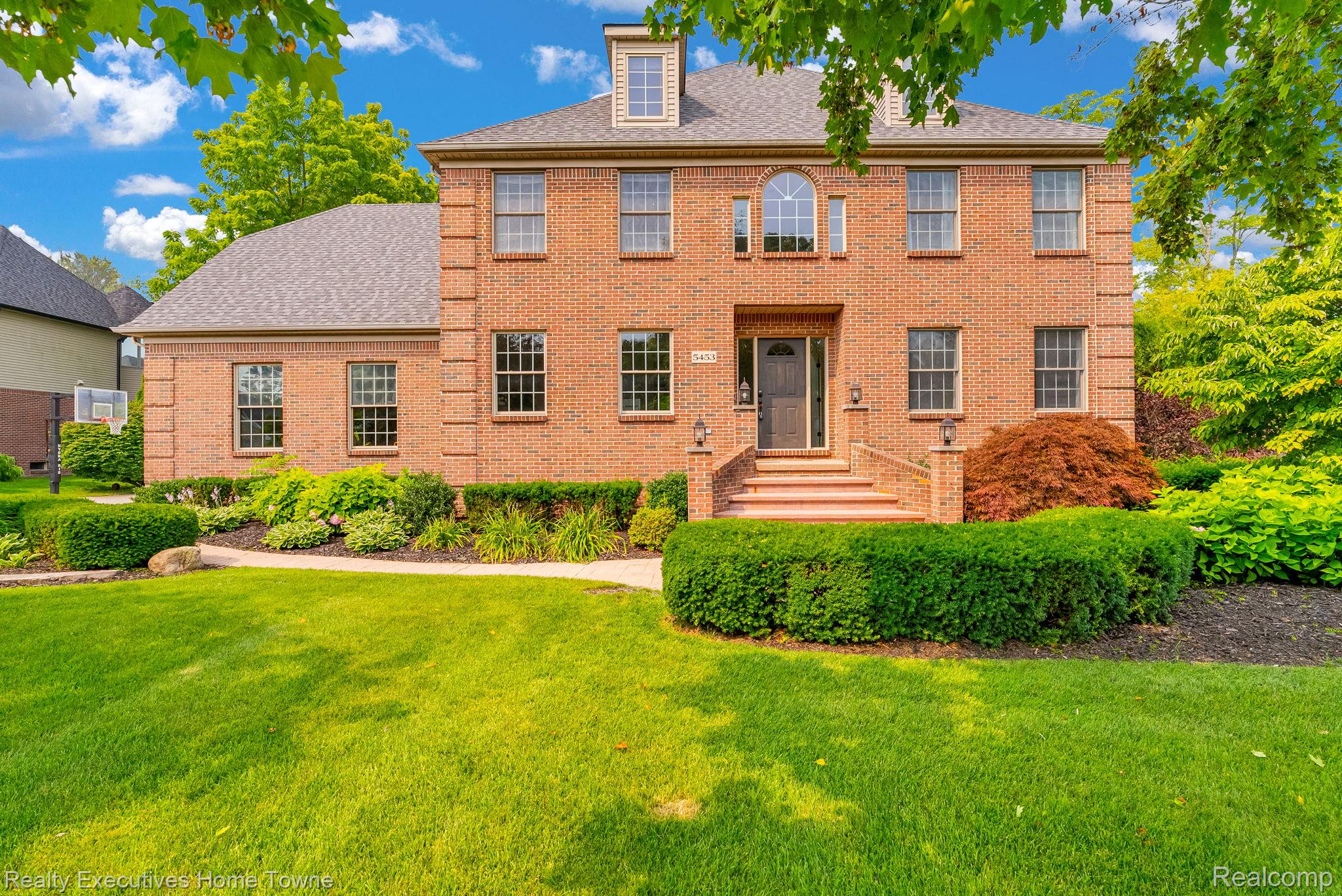 Two-story red brick house with an arched upper window, landscaped front garden, and broad lawn.