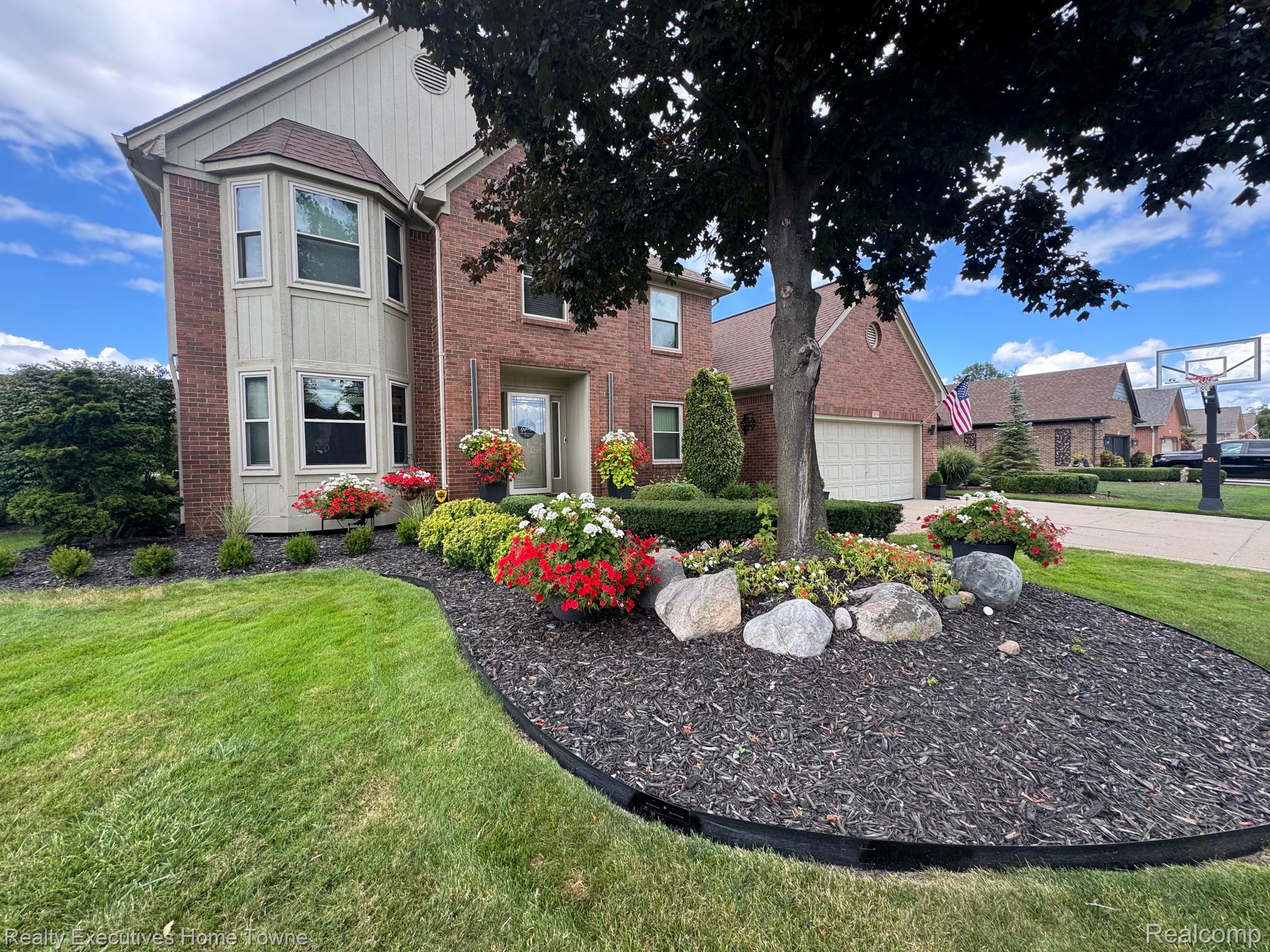 Two-story brick house with a bay window, flower beds, and a large shade tree.