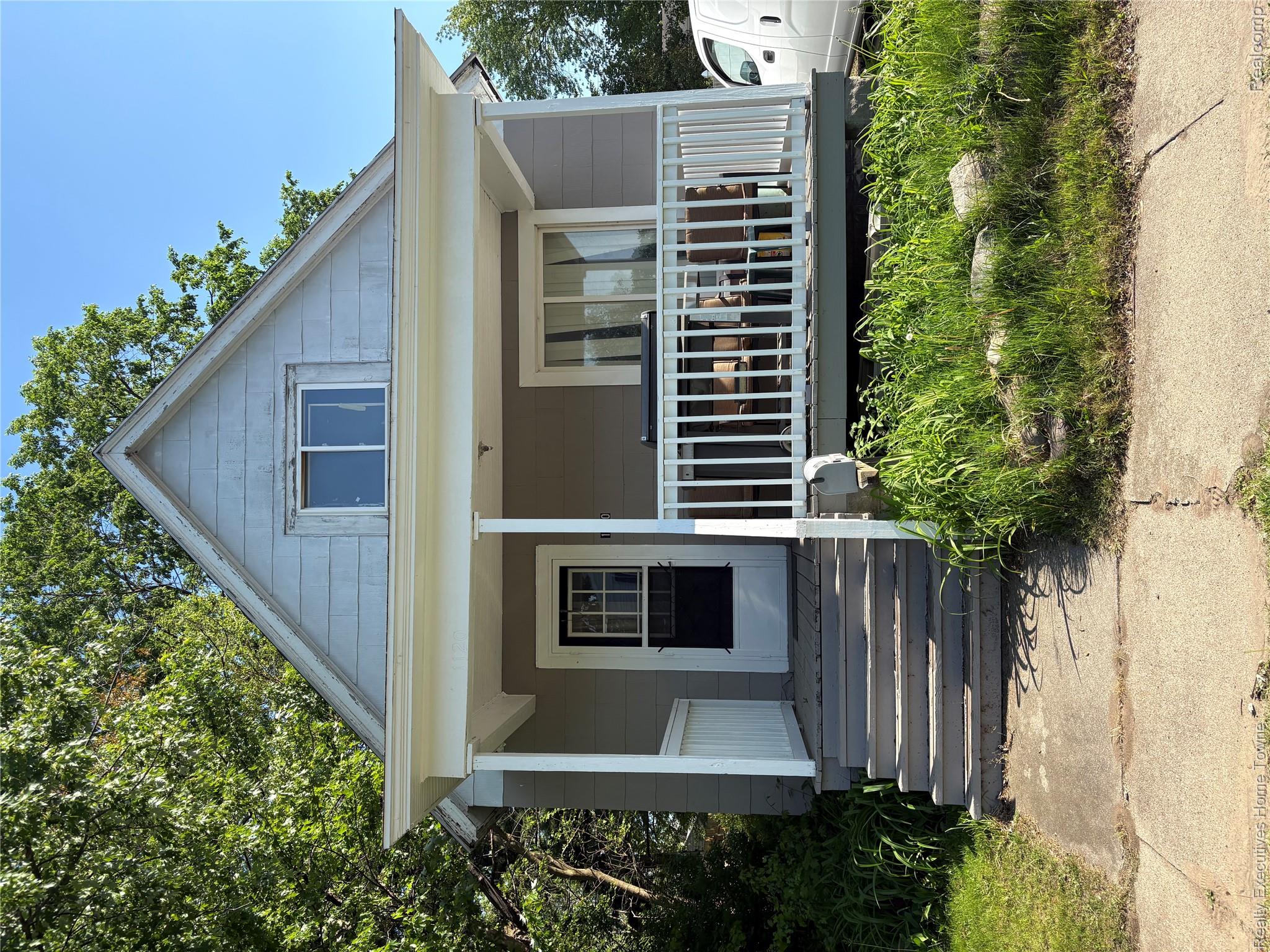 Gray gabled house with a covered porch, white railings, and steps leading to the sidewalk.