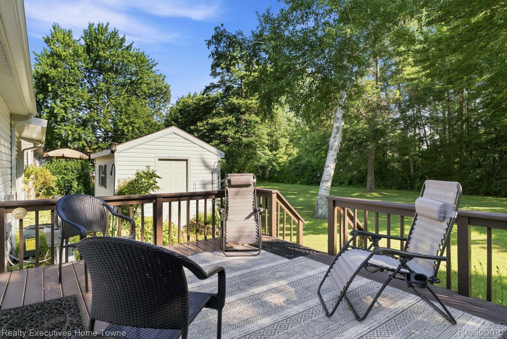 Furnished backyard deck with lounge chairs overlooking a leafy lawn and white storage shed.