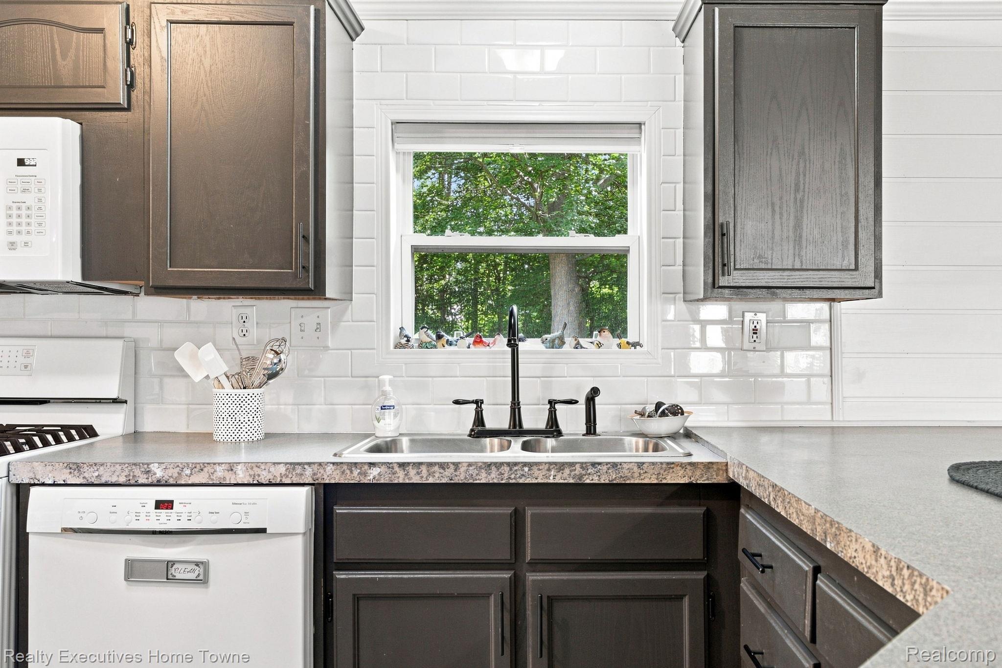 Kitchen with dark cabinets, white subway tile, and a sink beneath a window overlooking trees.