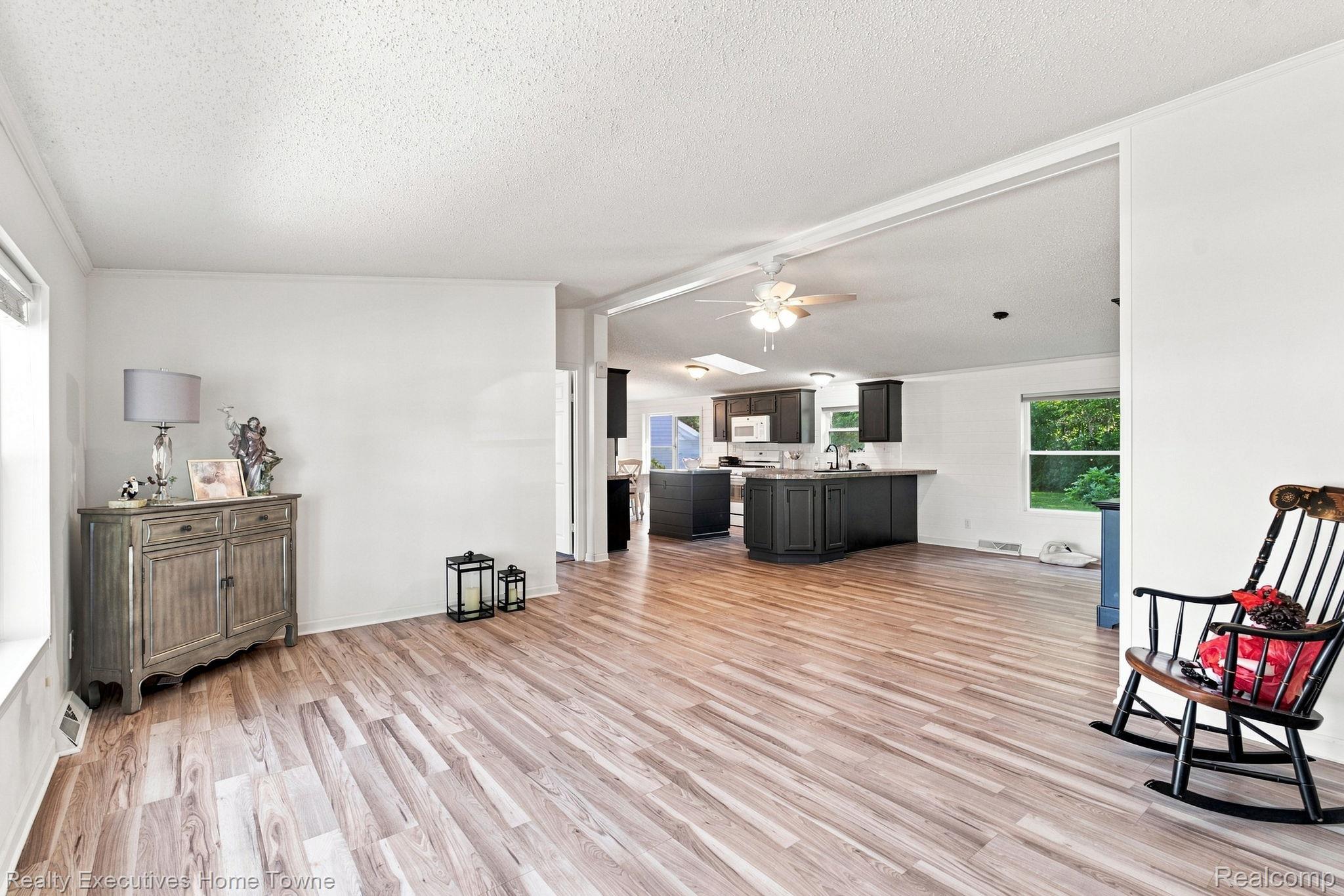 Open living room with wood-look flooring, black rocking chair, and dark-cabinet kitchen beyond.