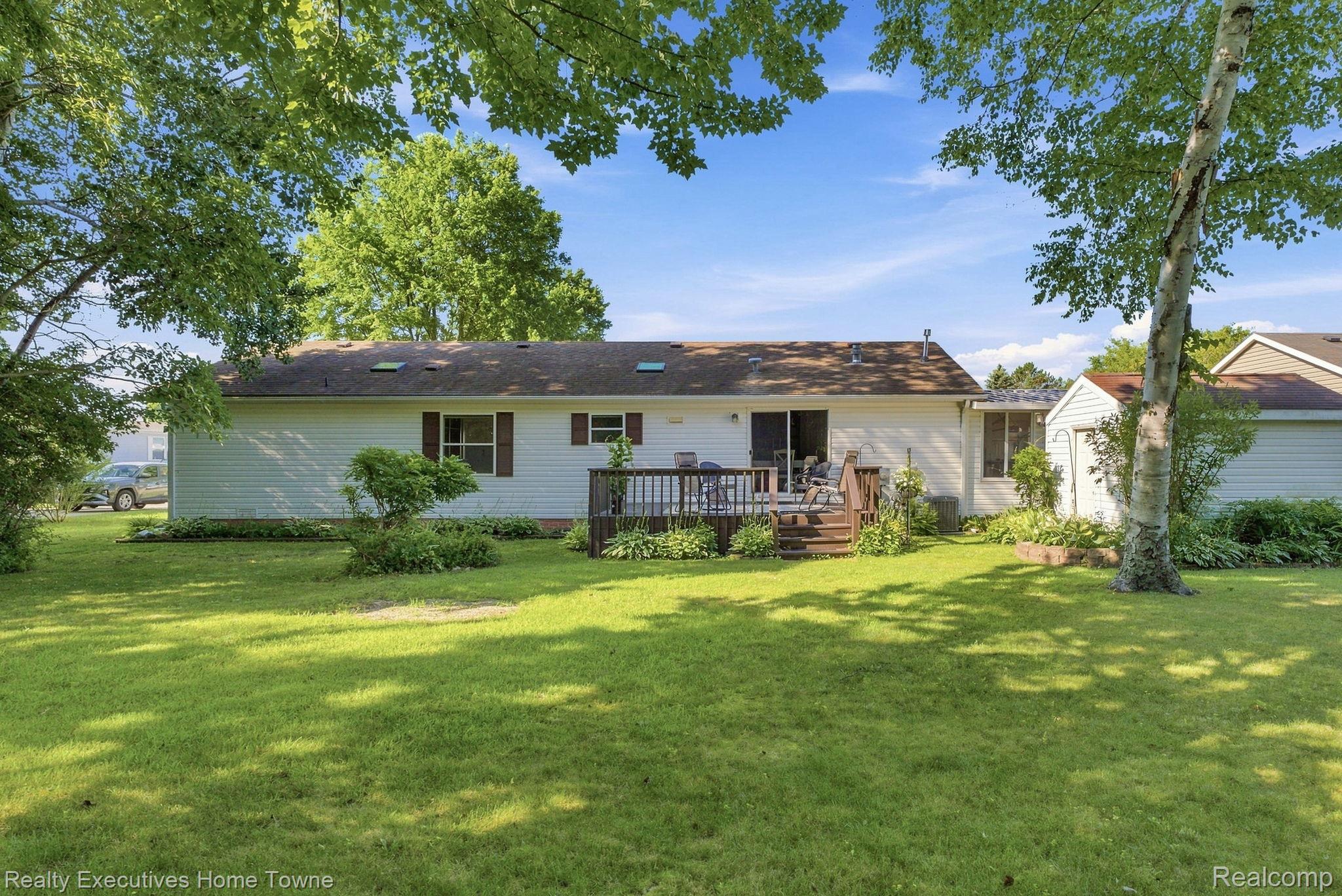 Single-story home with a wooden deck and patio chairs overlooking a broad, tree-shaded lawn.
