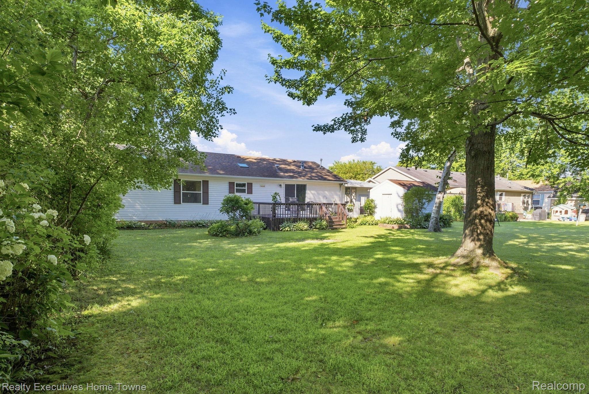 Rear of a single-story home with a wooden deck, expansive lawn, and mature shade trees.