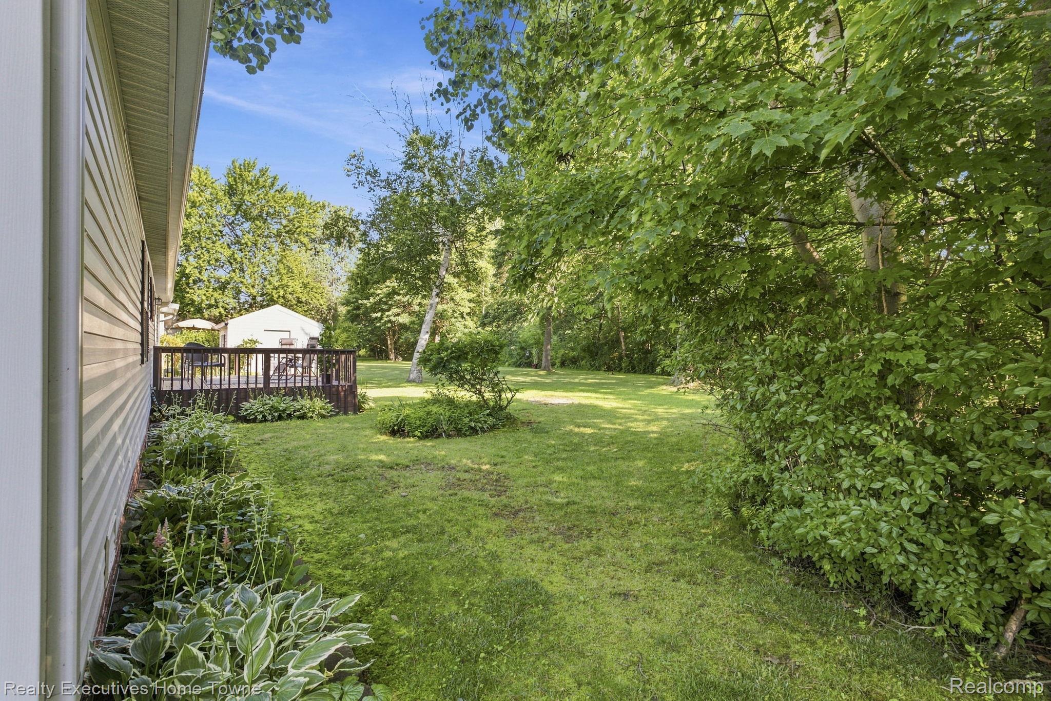 Backyard lawn and leafy trees beside the house, with garden beds and a raised deck.