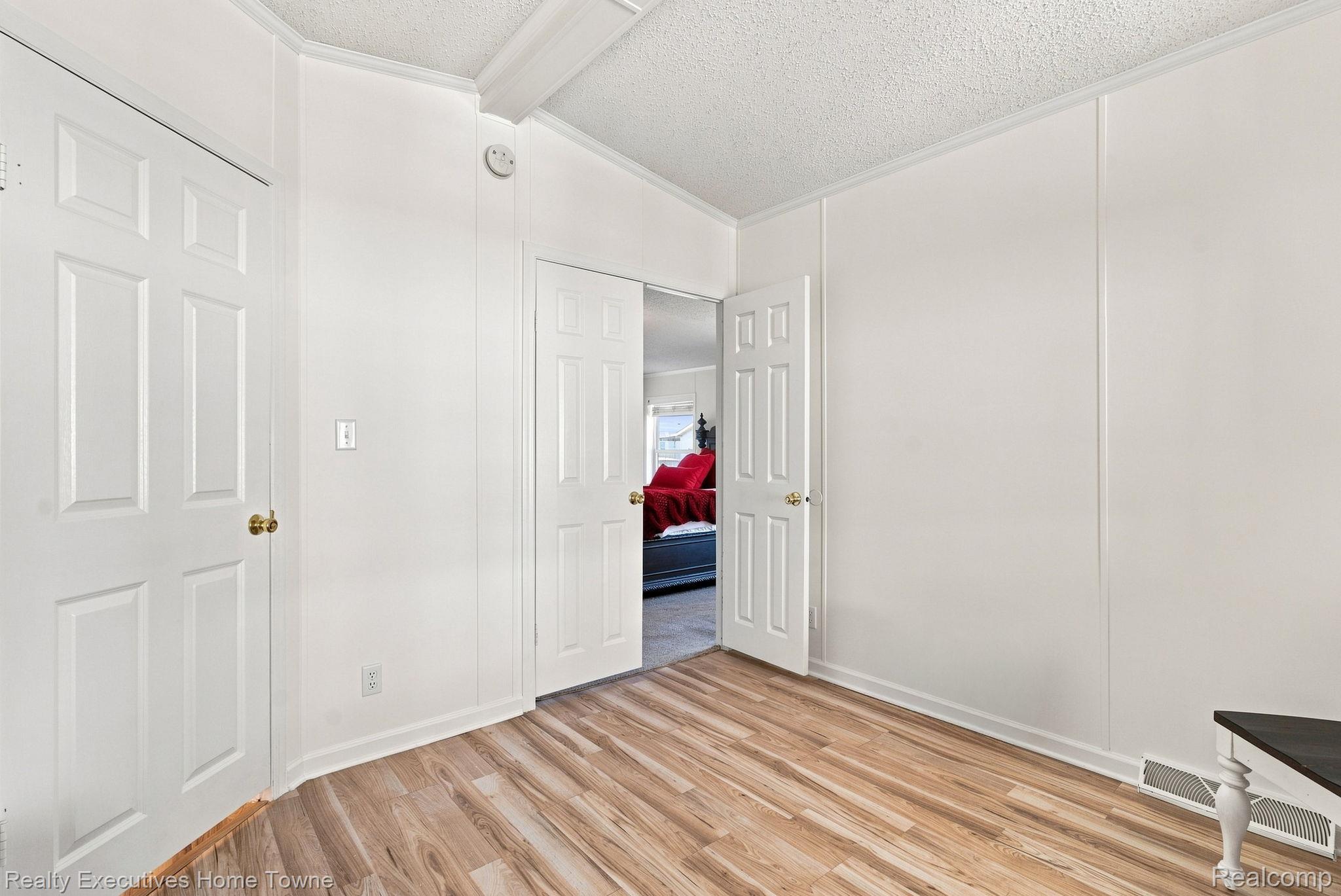 Mostly empty bedroom with wood-look flooring and open doors revealing a room with a red bed.