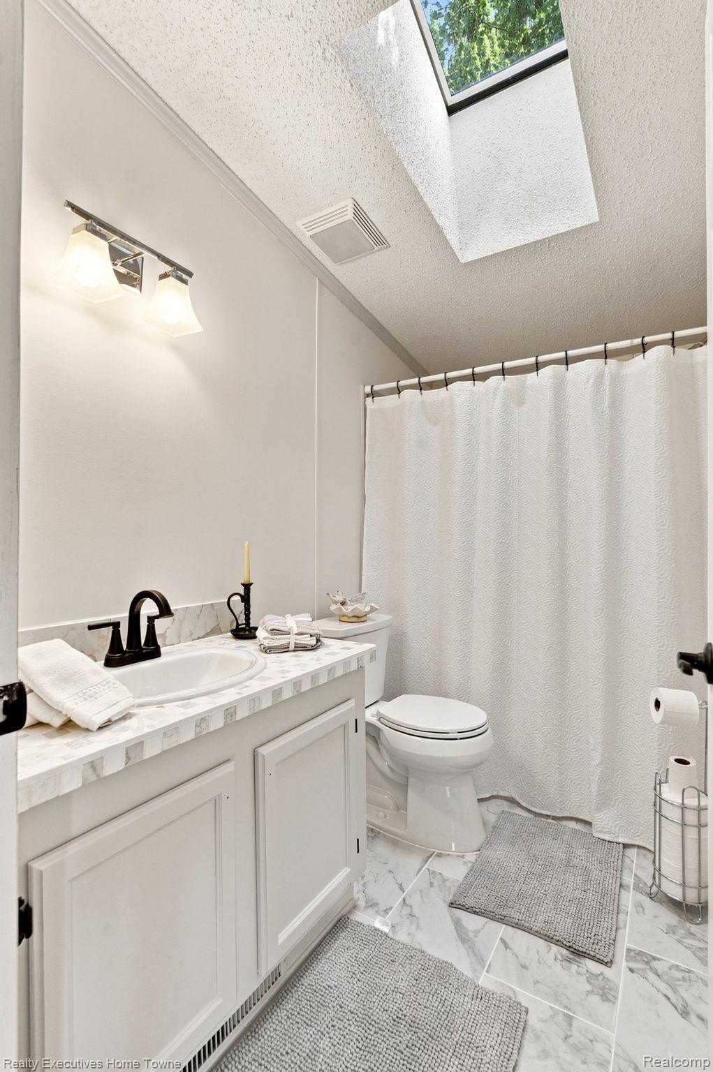 Bathroom with a white vanity, marble-look surfaces, and a skylight above the shower.