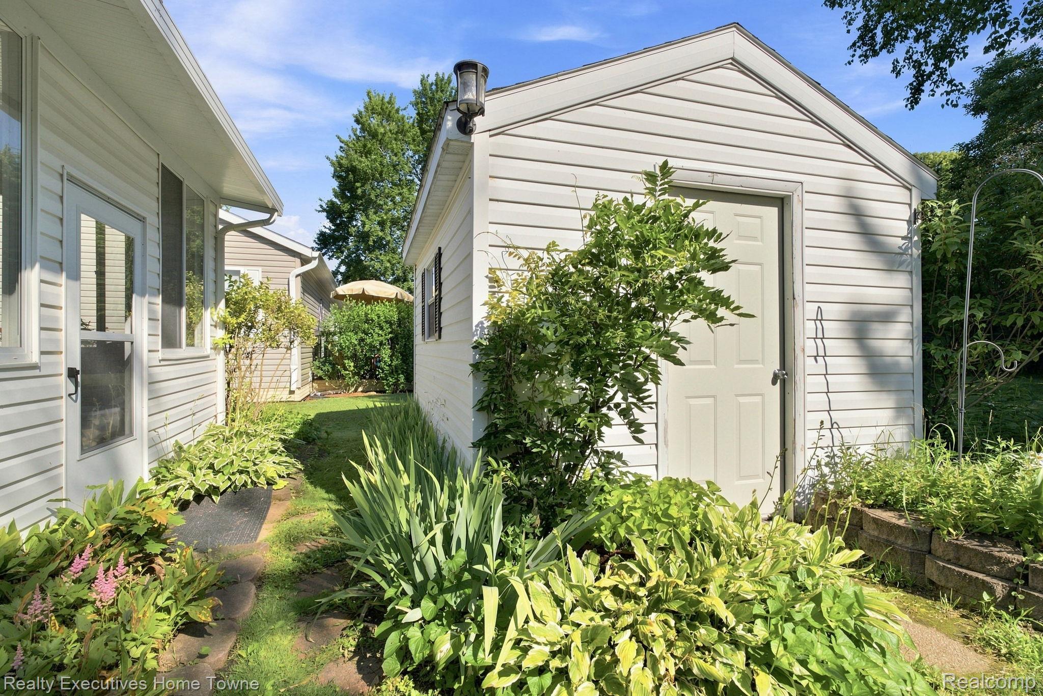 Side yard garden path between white-sided buildings, with dense greenery and a white outbuilding.