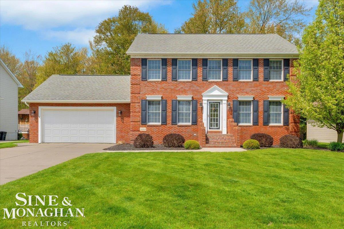 Two-story red brick house with dark shutters, an attached double garage, and a broad green lawn.
