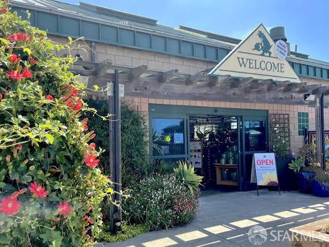 Storefront entrance beneath a wooden pergola, framed by flowering shrubs and marked with a WELCOME sign.
