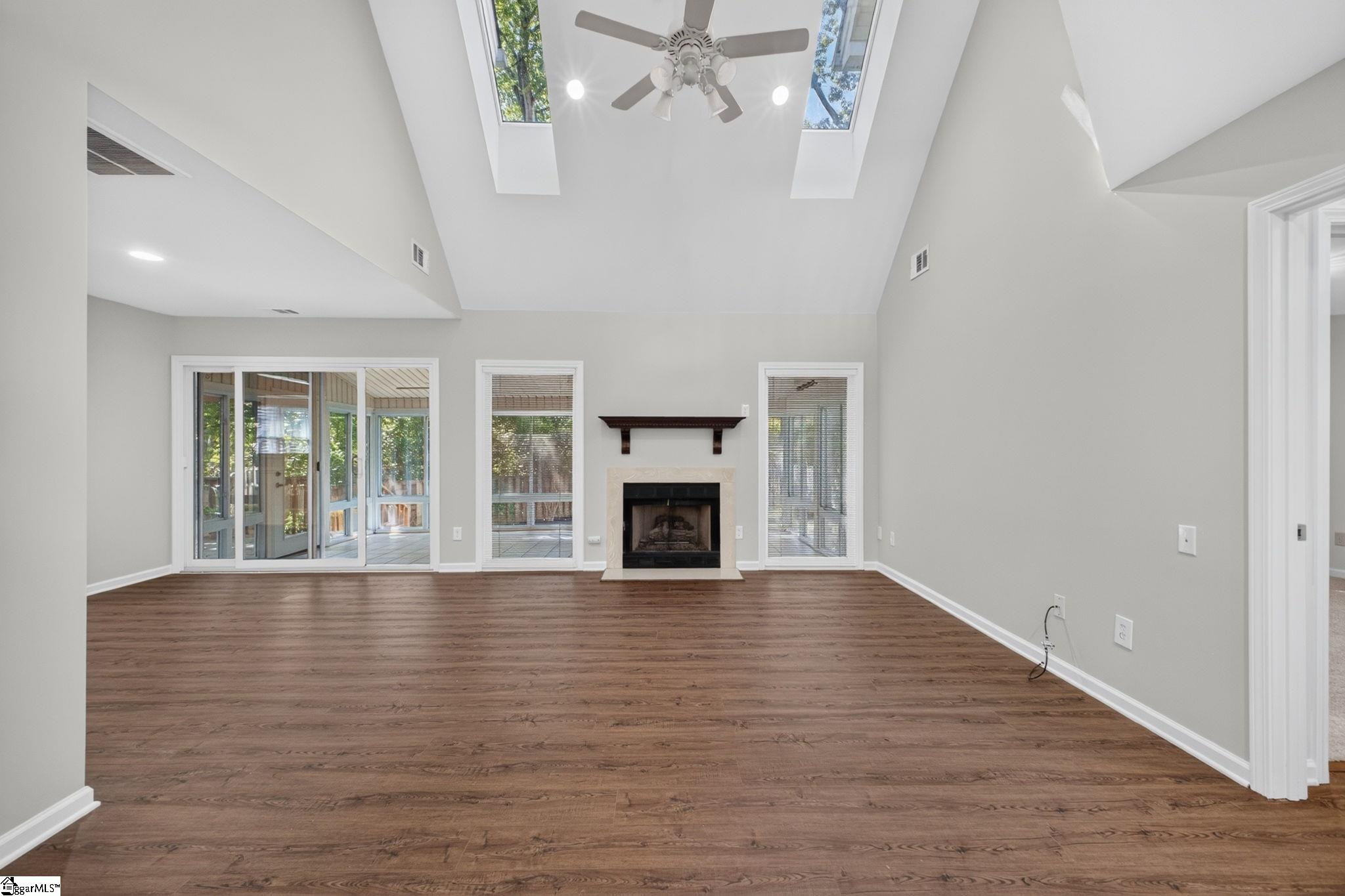 Empty living room with a fireplace, vaulted skylit ceiling, wood floors, and sliding glass doors to a screened porch.