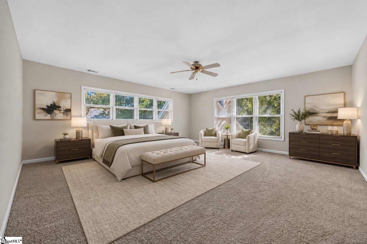 Staged primary bedroom with a king bed, window-side armchairs, and large windows overlooking greenery.