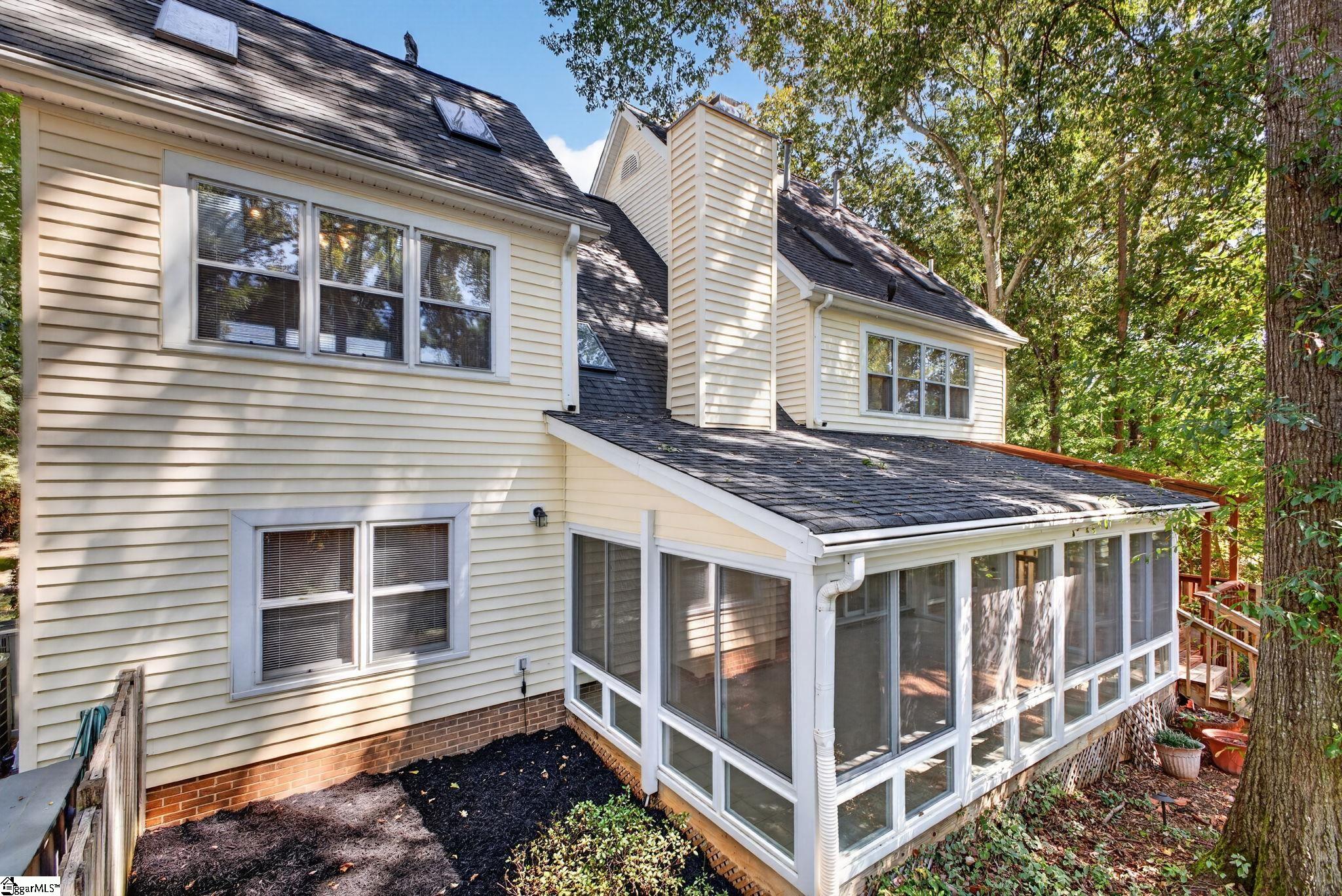 Rear exterior of a cream-sided home with a large glass-enclosed sunroom and mature trees.
