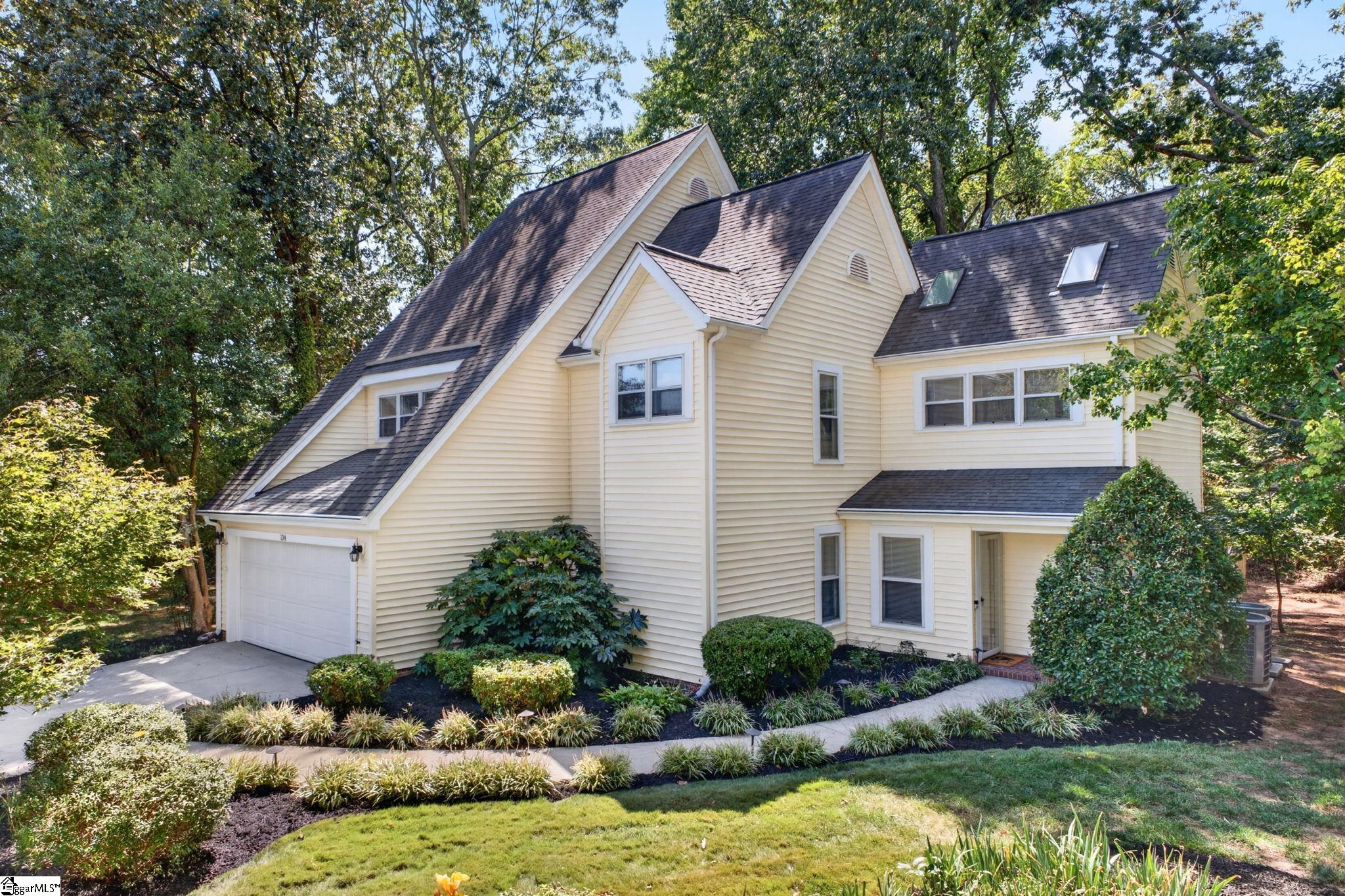 Cream-colored house with steep gabled roofs, an attached garage, and landscaped garden beds among mature trees.