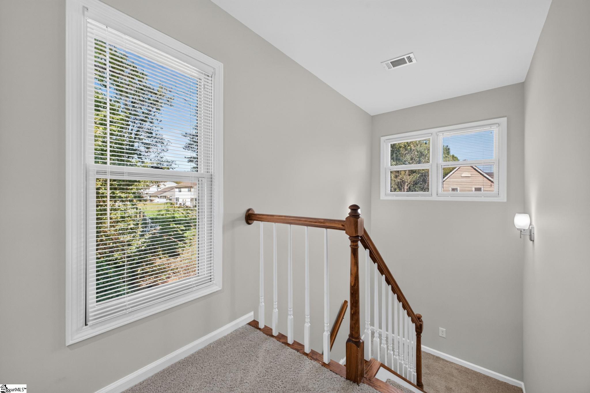 Upstairs stair landing with gray carpet, large windows, and a wooden handrail with white balusters.