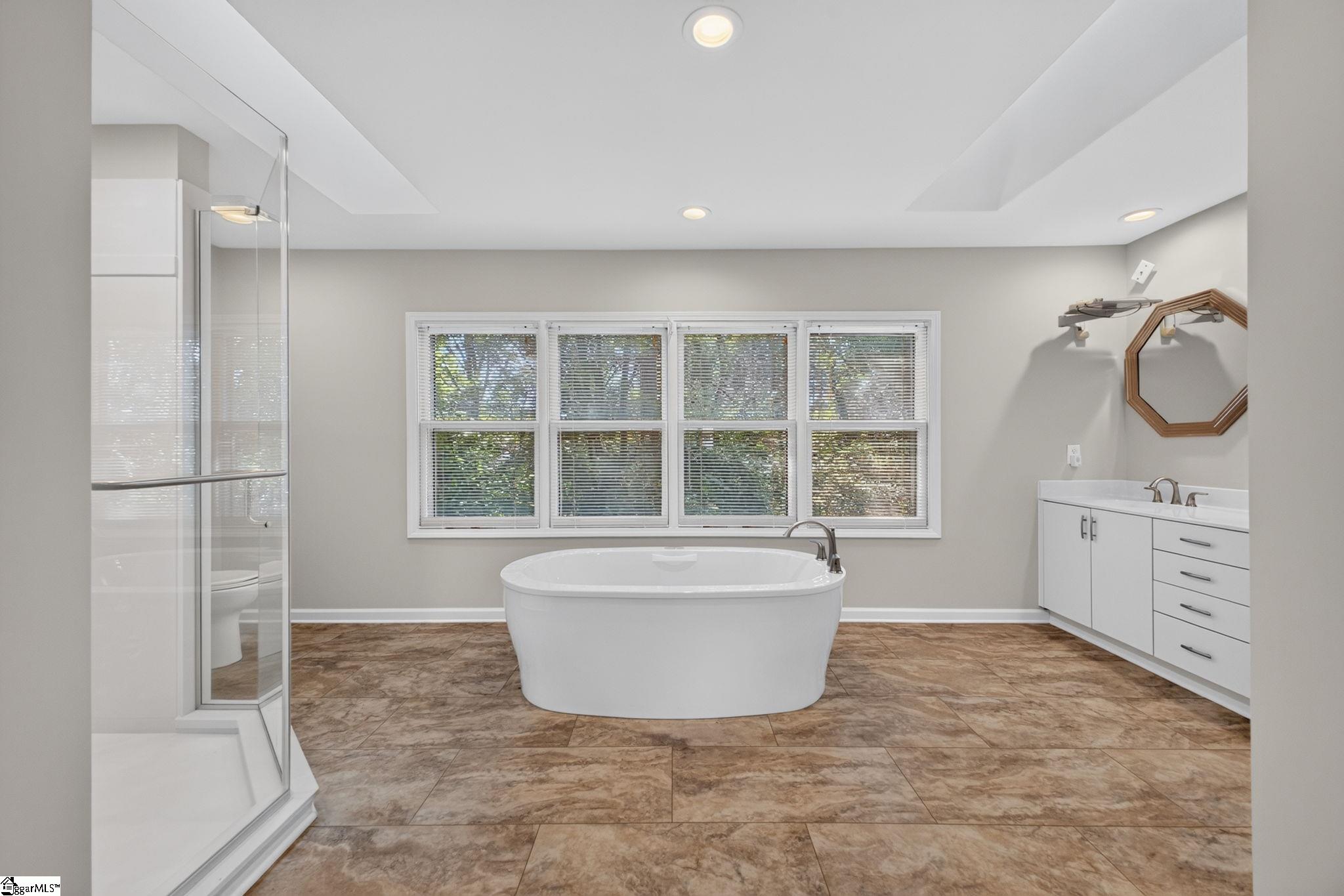 Bathroom with a freestanding tub beneath a wide window, glass shower, and white vanity.