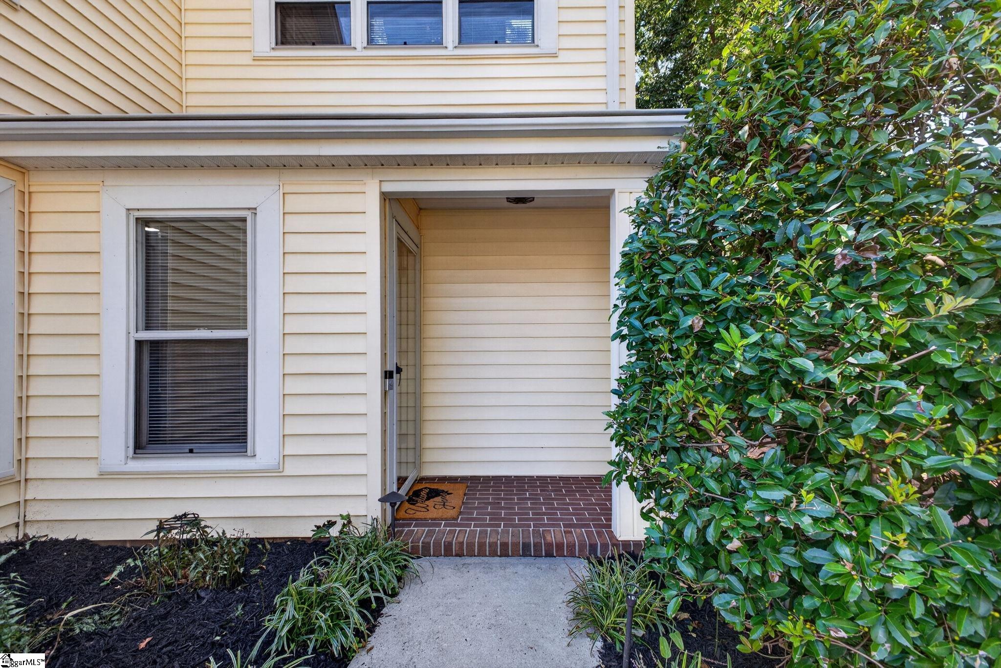 Cream-sided home entrance with a covered brick porch and dense shrubs.