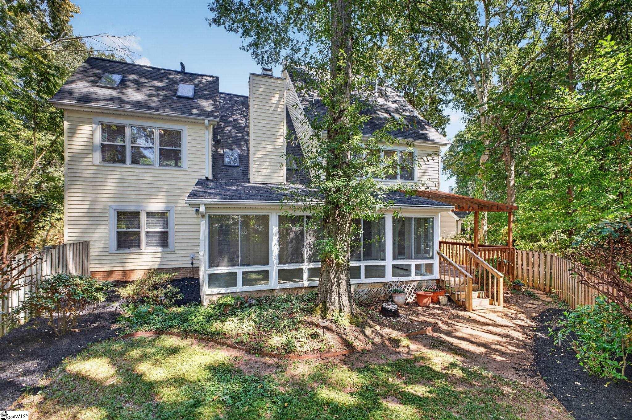 Rear of a beige two-story home with a glass-enclosed sunroom and wooden deck in a wooded yard.