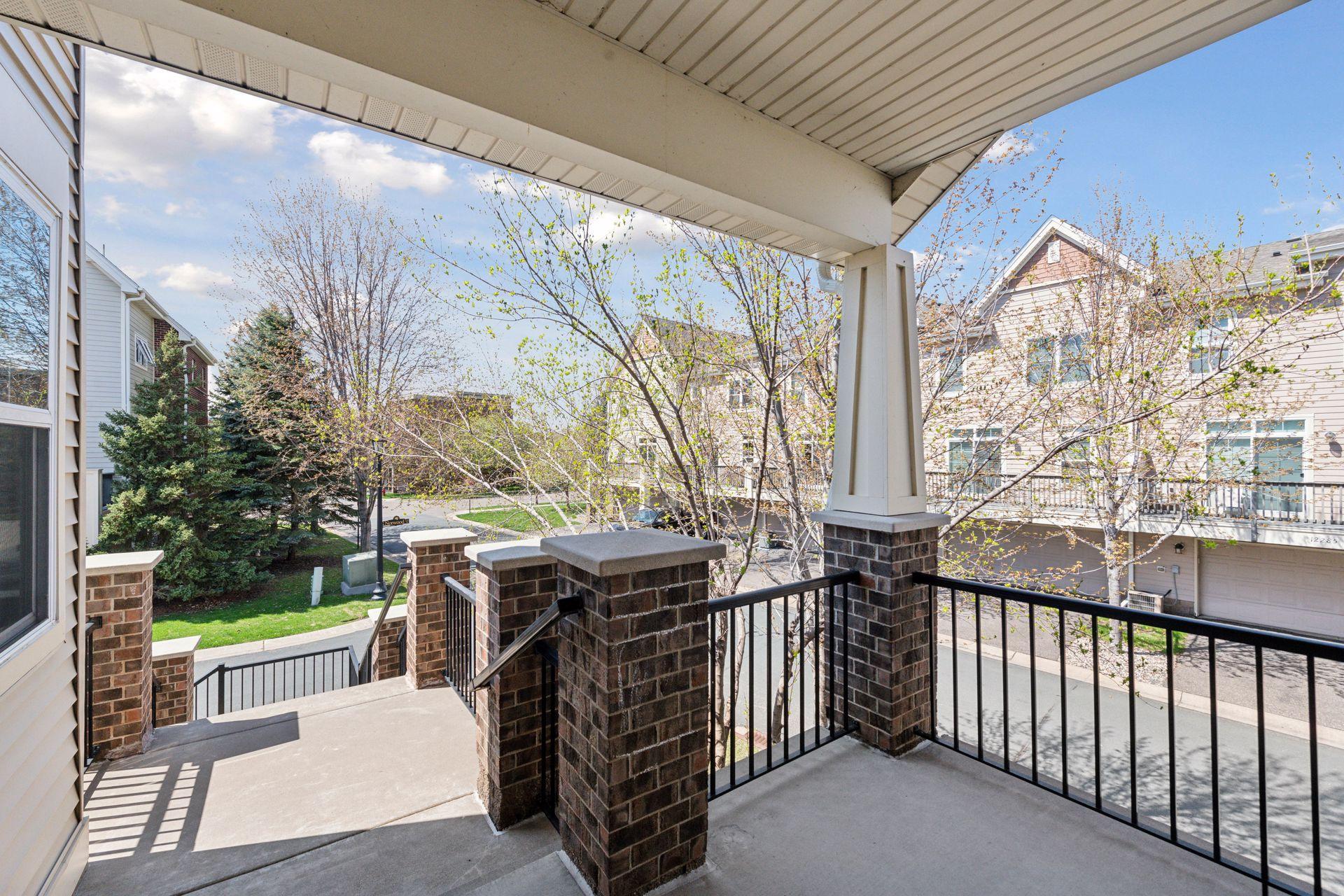 Covered balcony with brick columns and black railings overlooking neighboring homes and budding trees.