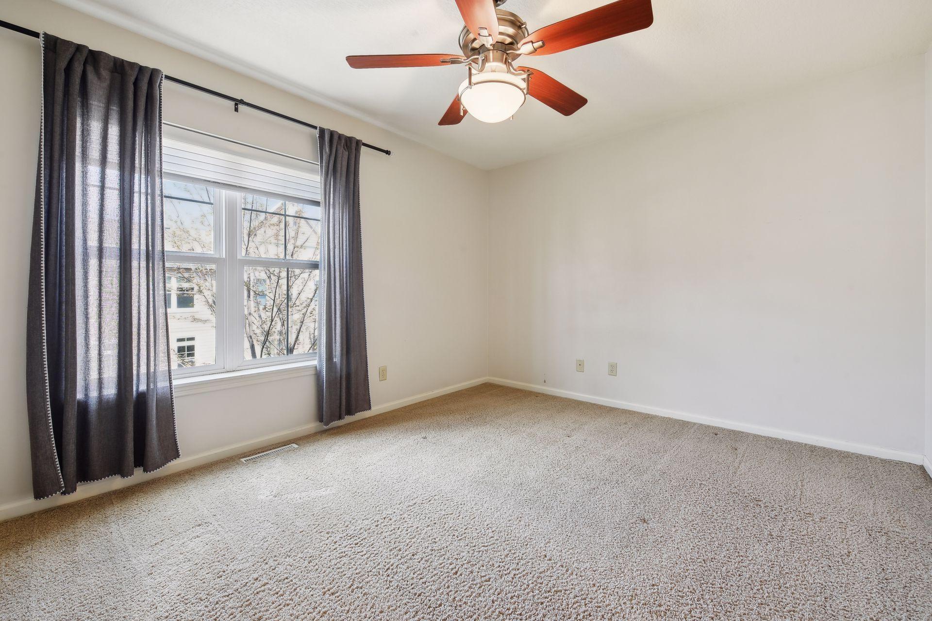 Empty carpeted bedroom with dark curtains, a large window, and a ceiling fan.