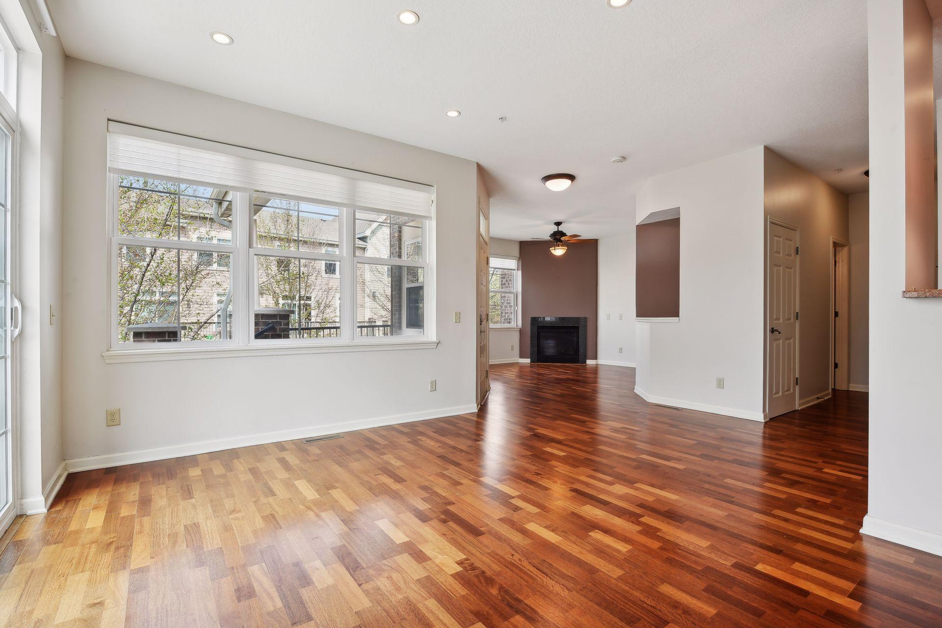 Empty living room with polished wood floors, large windows, and a fireplace.