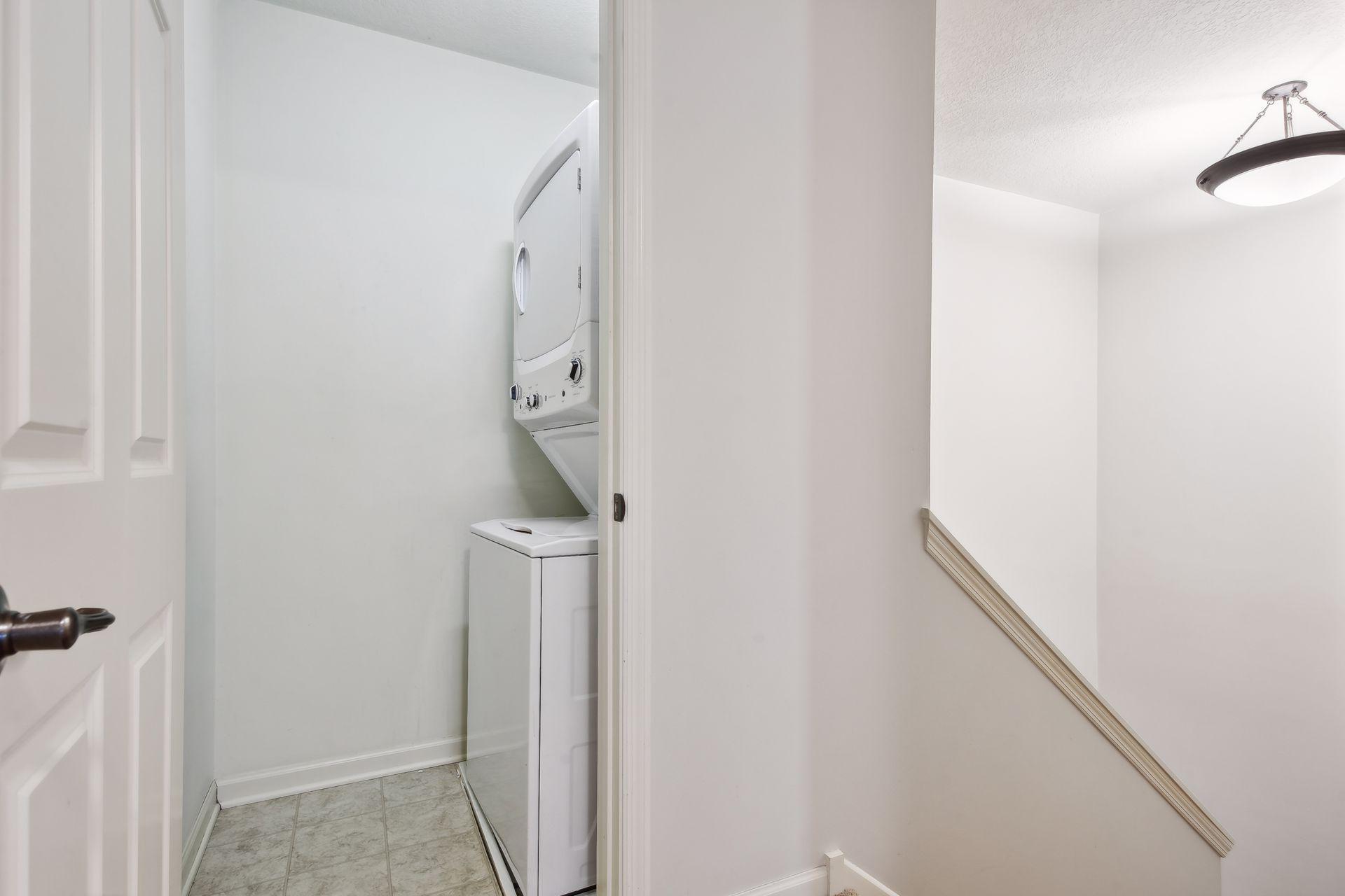 Laundry closet with a stacked washer and dryer beside a white stairwell.