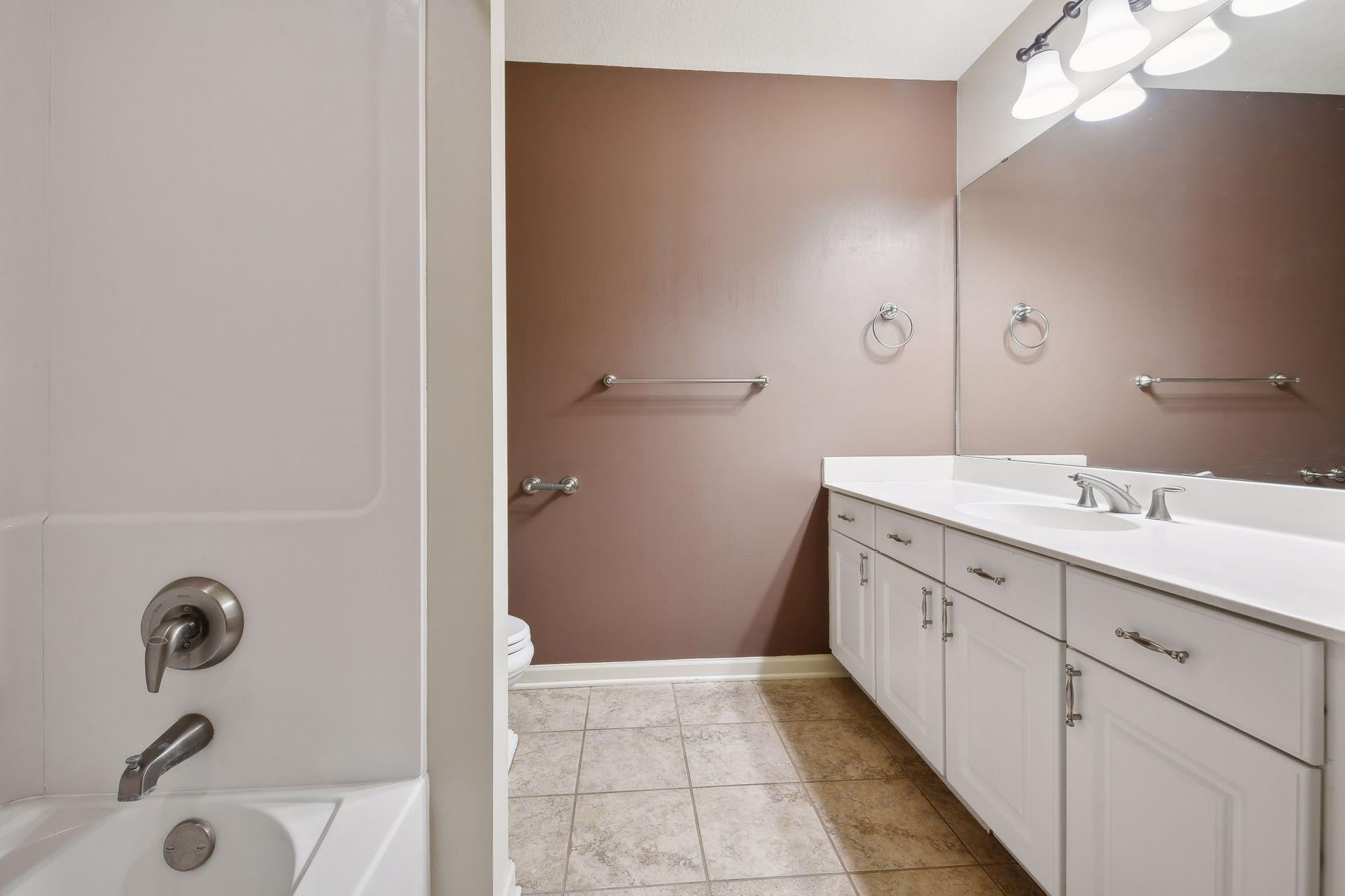 Bathroom with a white tub and shower, long vanity, large mirror, and chrome grab bars.