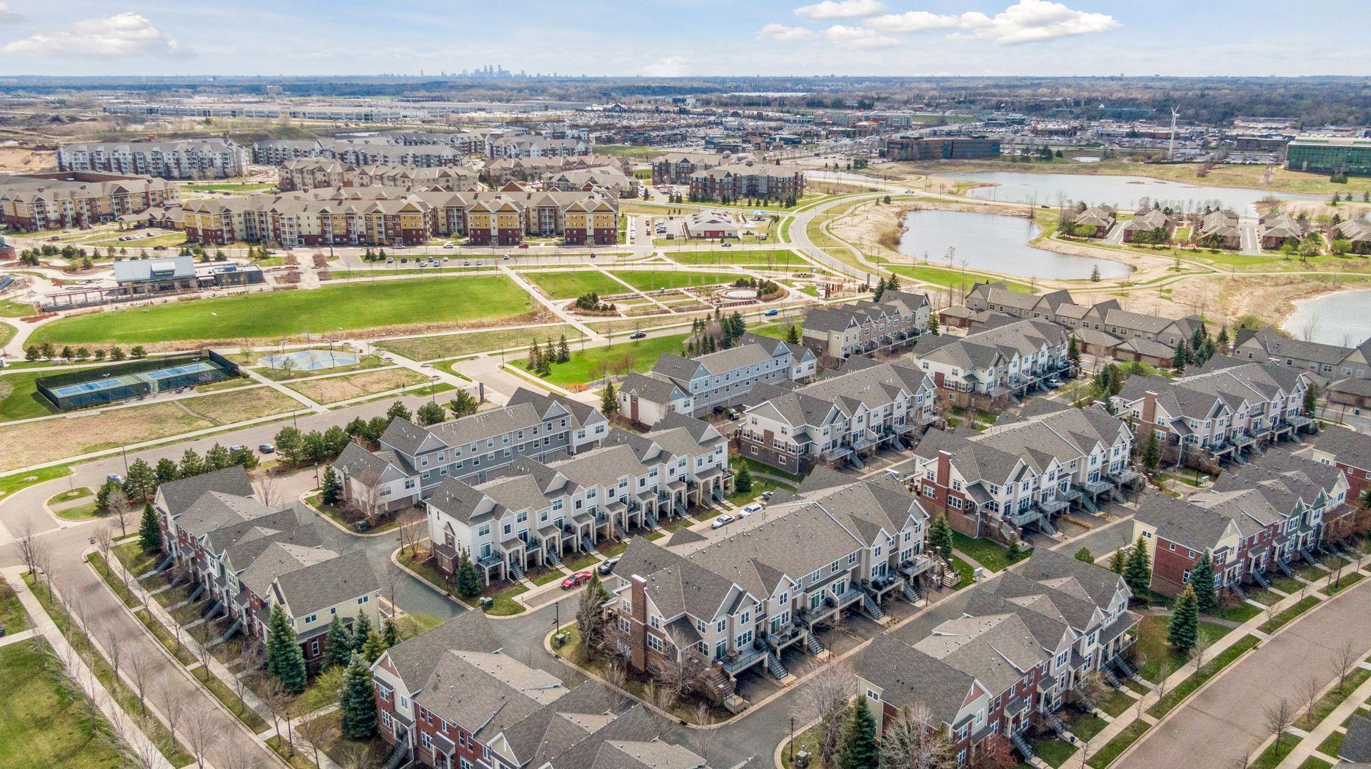 Aerial view of a townhouse neighborhood with ponds, tennis courts, and a distant city skyline.