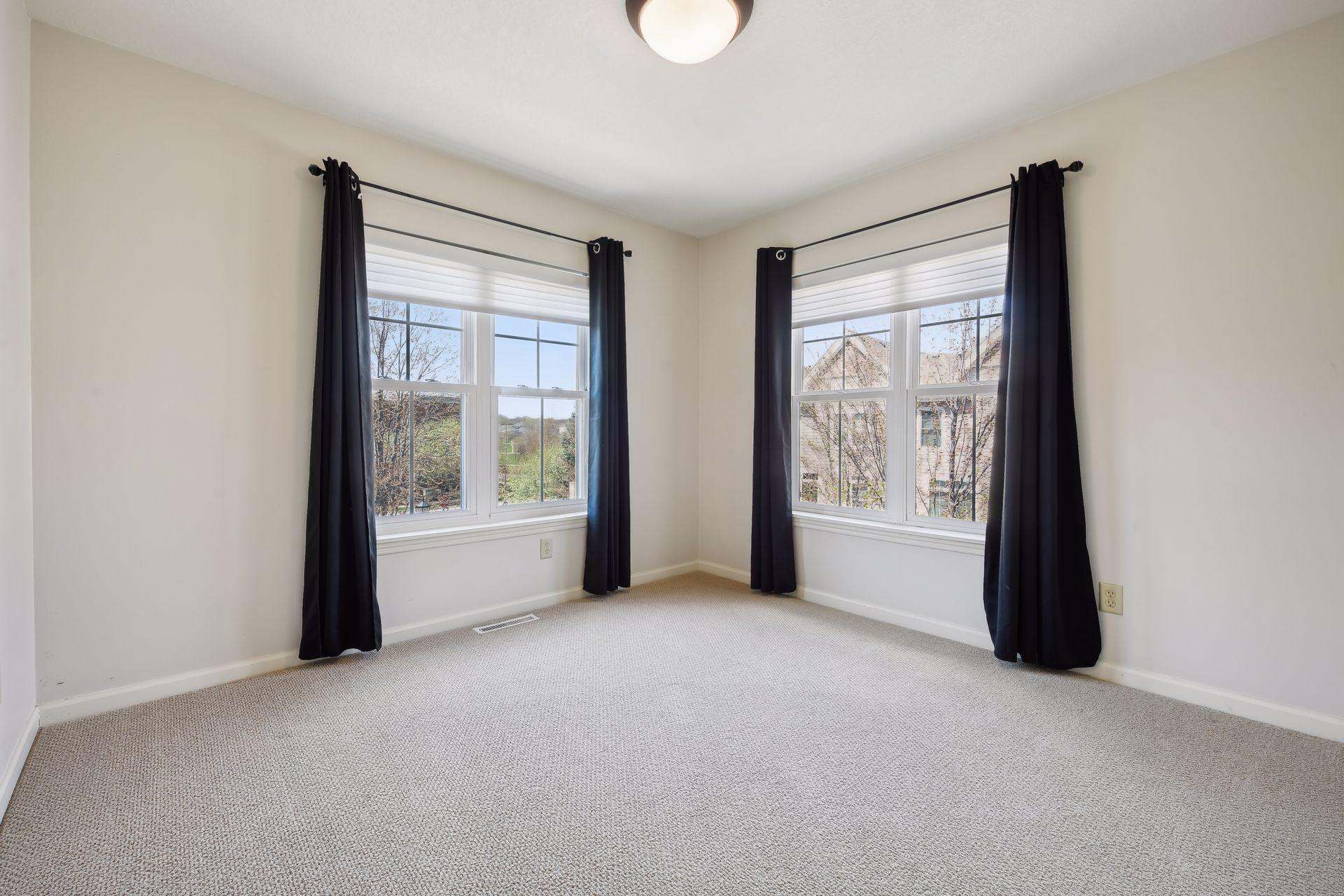 Empty carpeted room with two large windows framed by dark curtains.