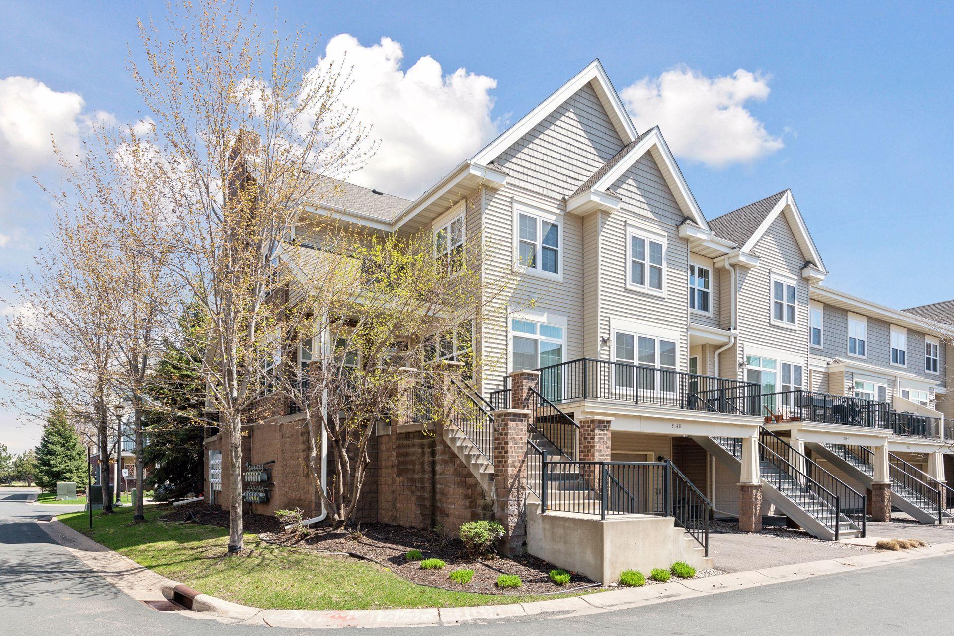 Attached beige townhome with peaked gables, raised black-railed decks, and a budding tree beside the street.