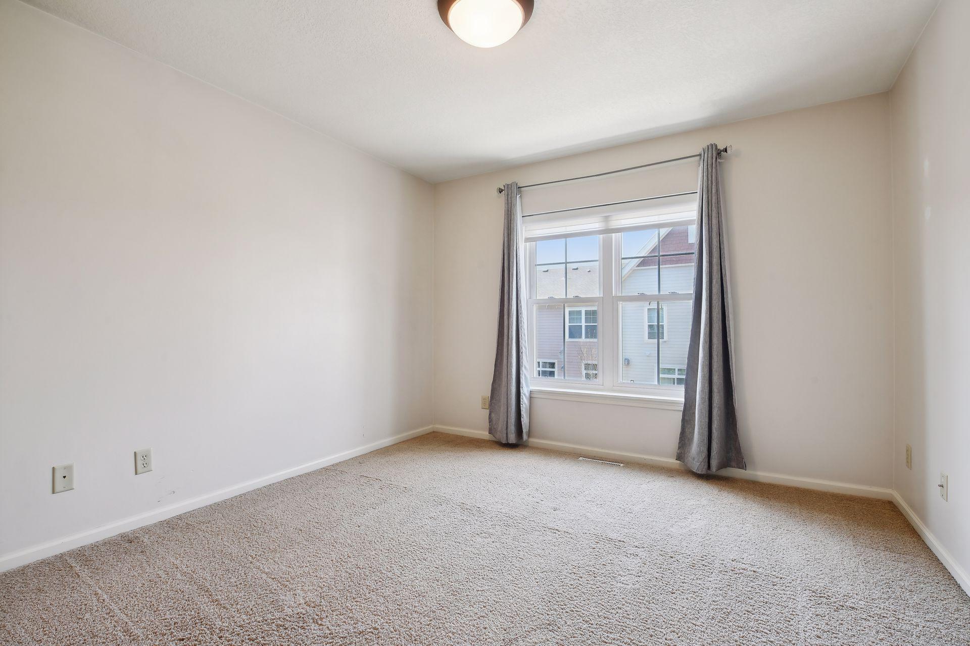 Empty carpeted bedroom with gray curtains framing a large window.