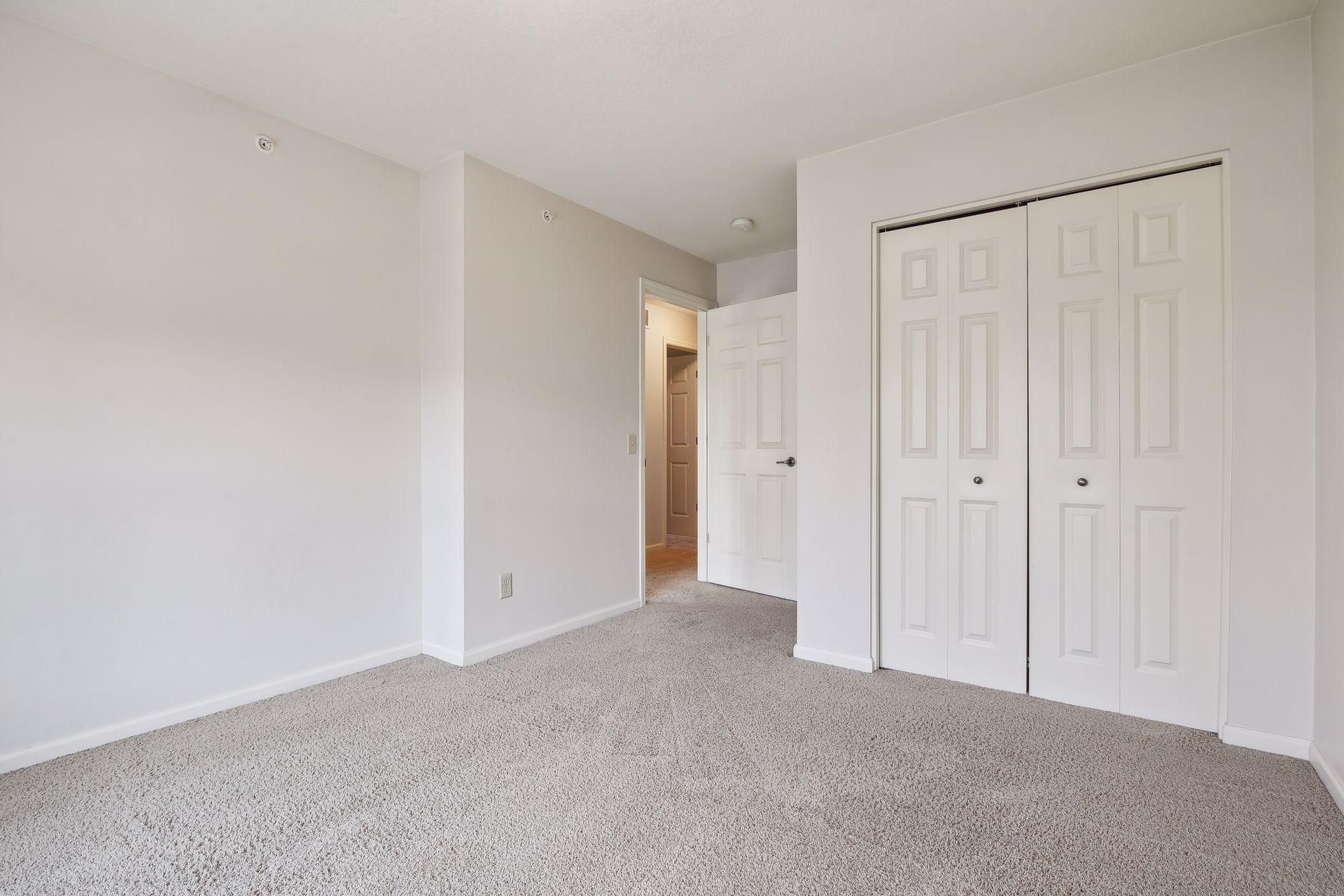 Empty bedroom with beige carpet, a double-door closet, and an open doorway to the hall.