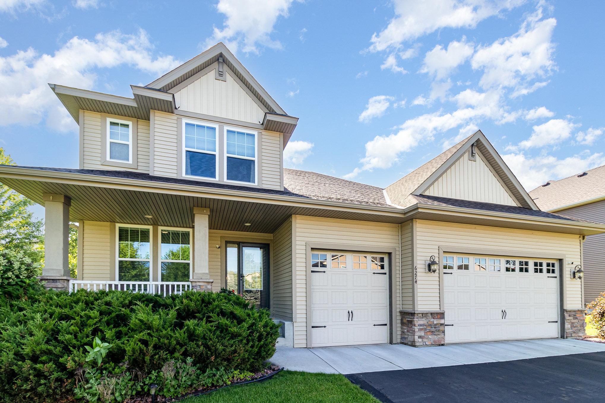 Two-story beige home with a covered porch, landscaped shrubs, and a wide two-car garage.