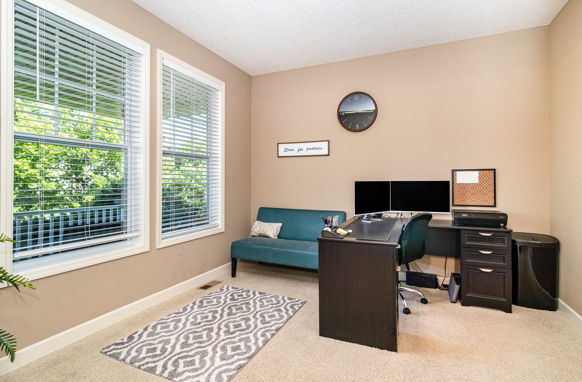 Home office with a dark L-shaped desk, dual monitors, teal loveseat, and two large windows.