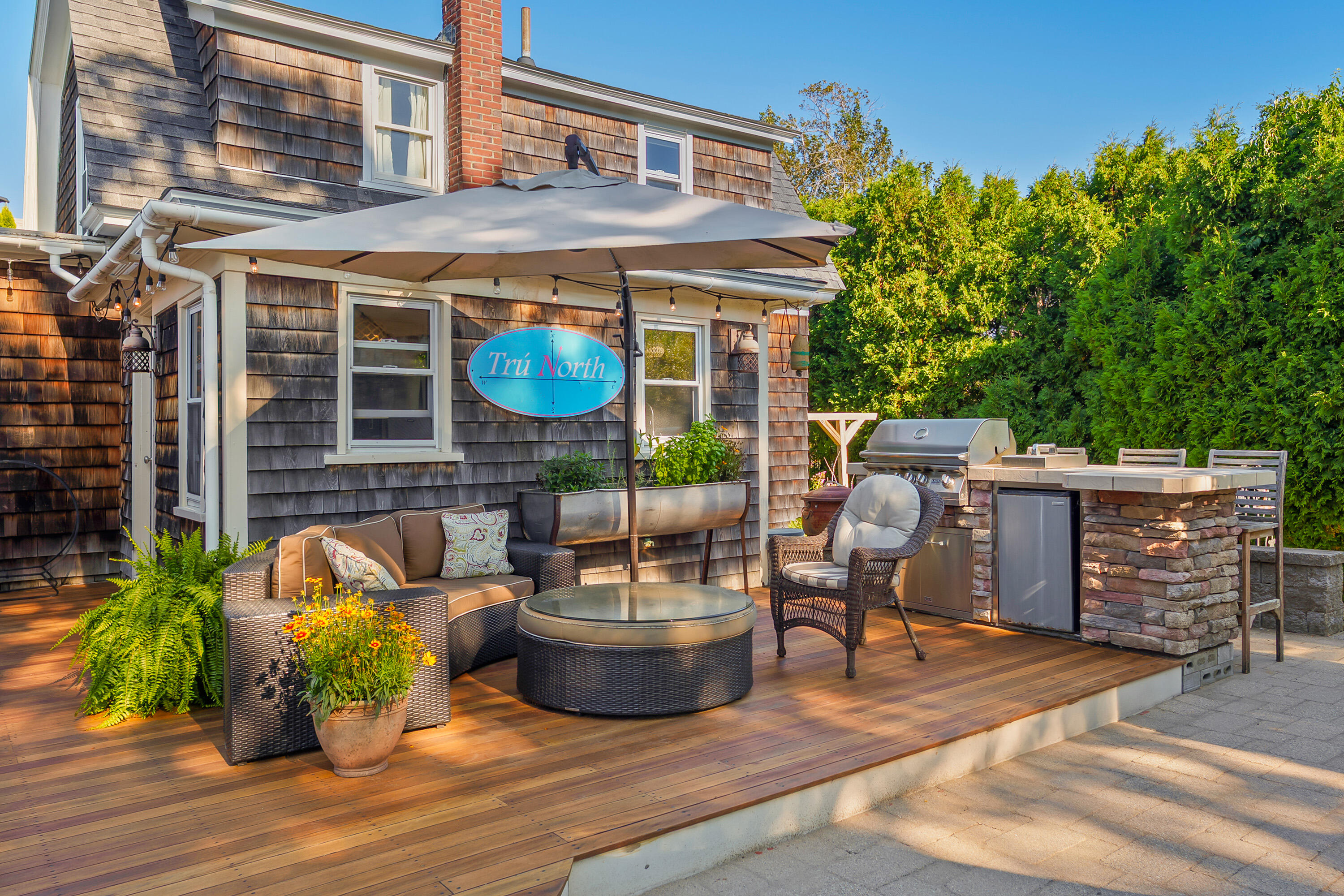 Furnished wooden deck with wicker seating, a shade umbrella, potted flowers, and an outdoor grill.