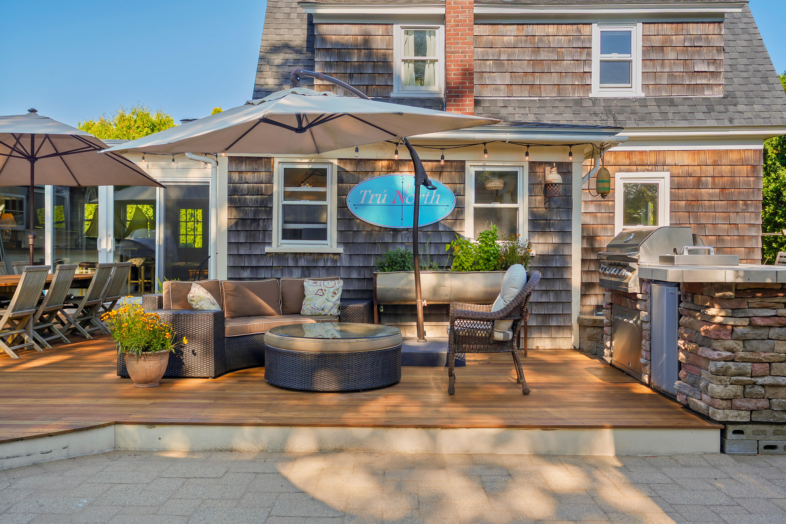 Outdoor deck with wicker seating beneath umbrellas, string lights, and a stone-base grill.