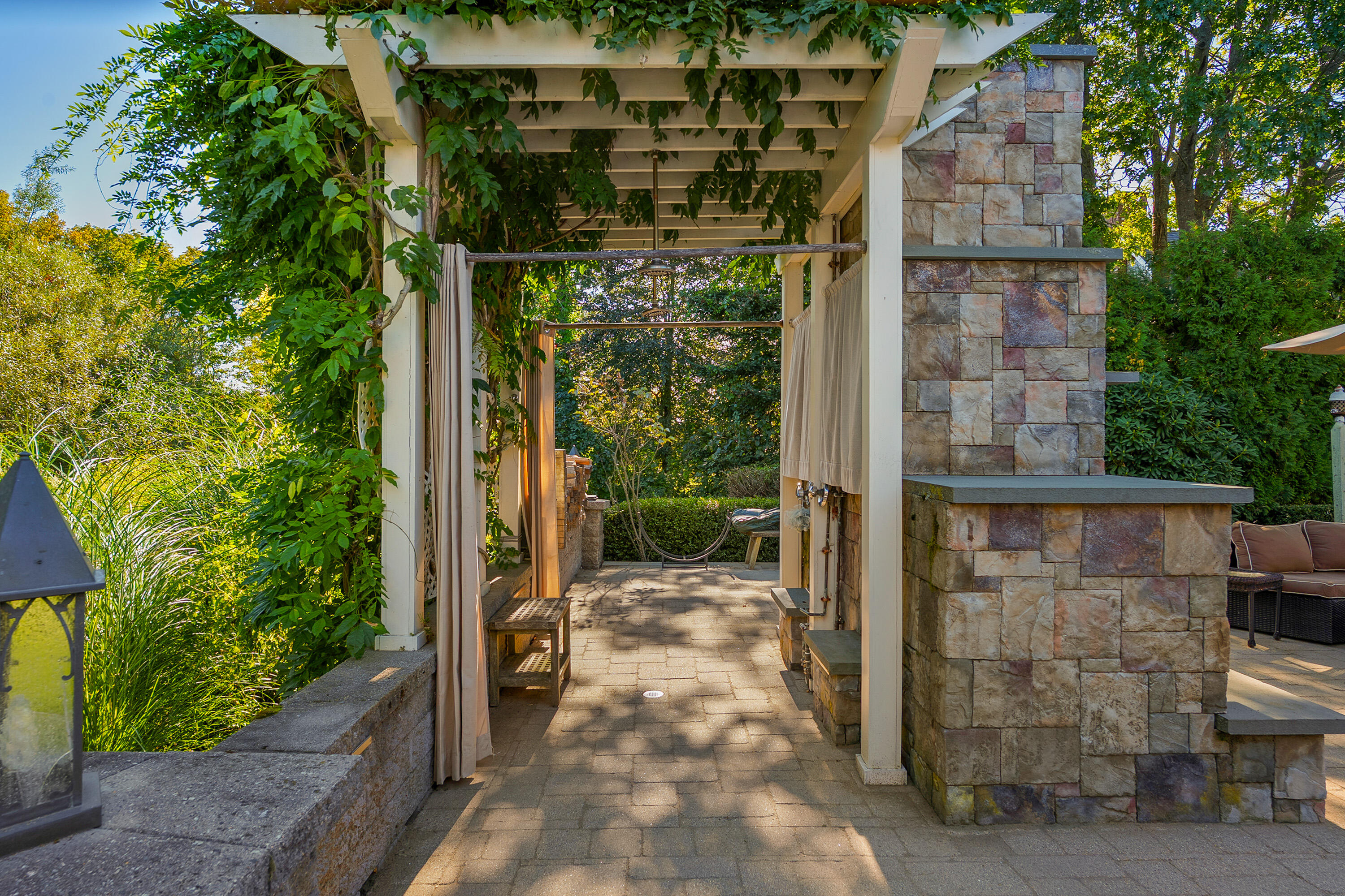 Garden patio beneath a vine-covered pergola, with a stone fireplace and hammock.