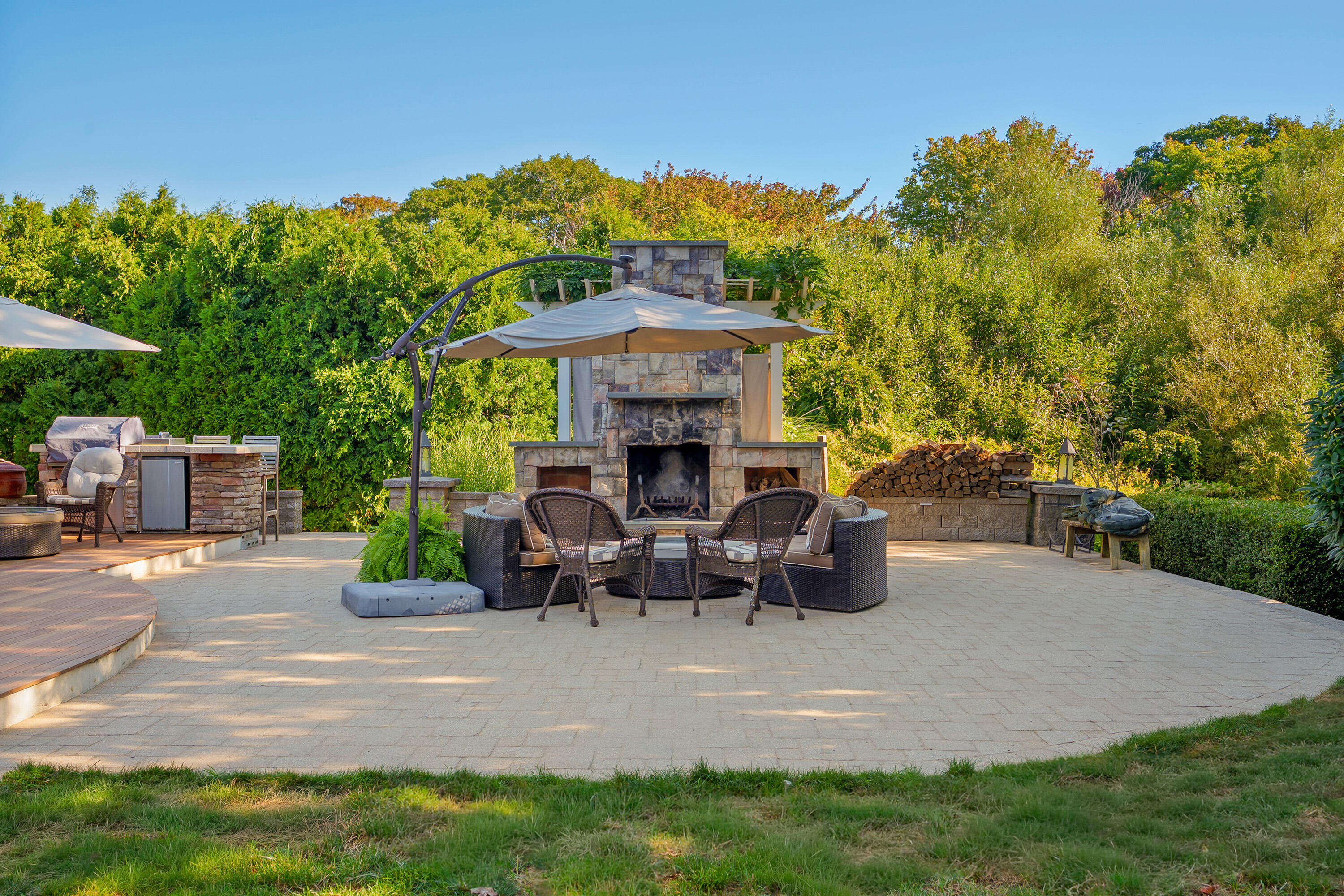 Outdoor patio with a stone fireplace, umbrella-shaded seating, built-in grill, and stacked firewood.