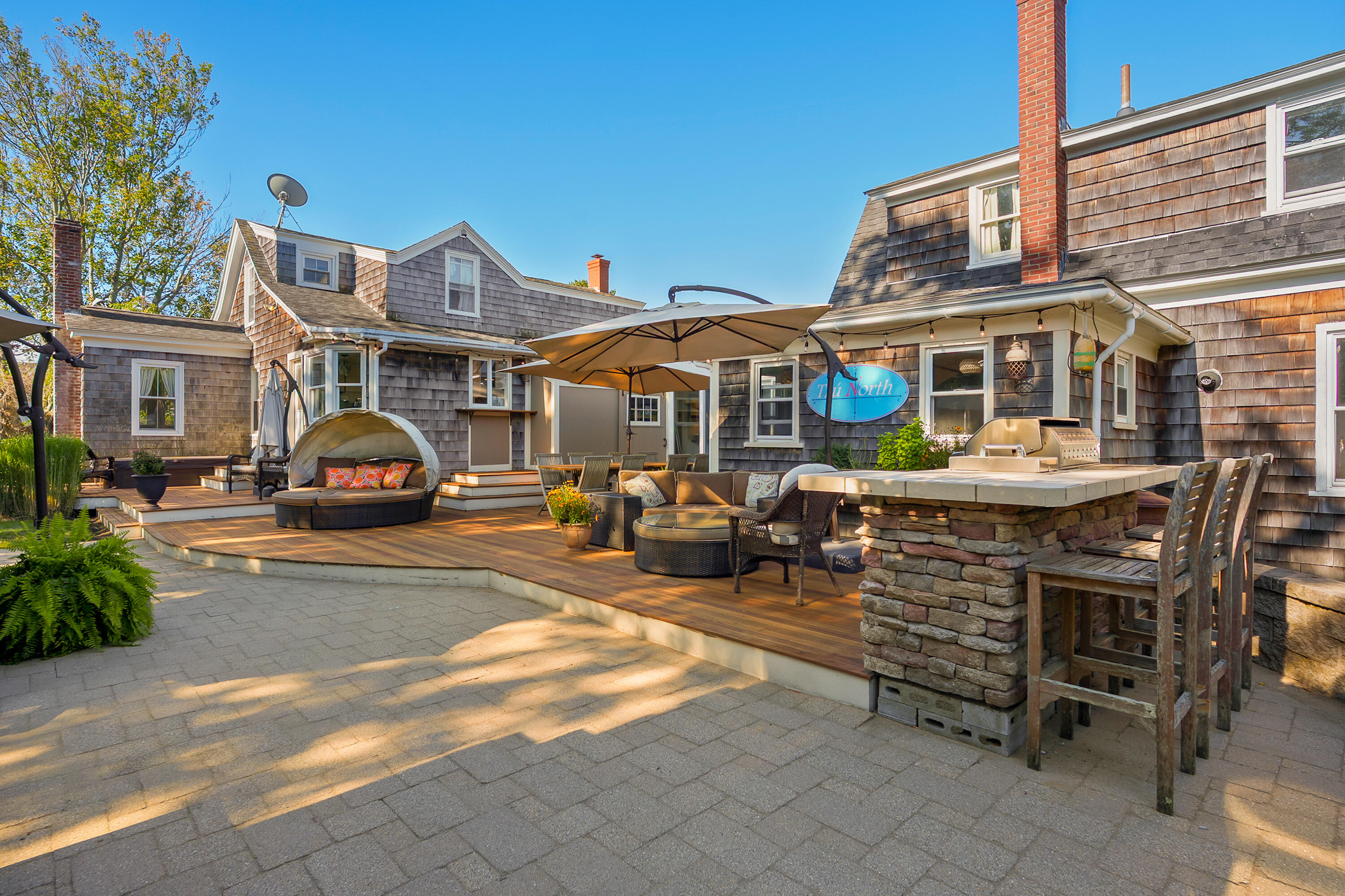 Shingle-sided home with a furnished wood deck, stone outdoor kitchen, grill, and umbrella-shaded seating.