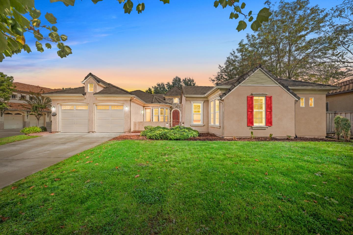 Single-story stucco home with a two-car garage, red shutters, and a broad front lawn at dusk.