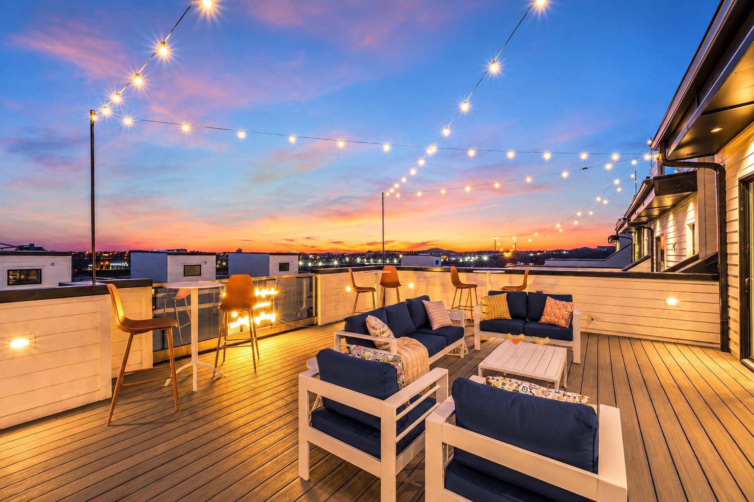 Rooftop deck with lounge seating and string lights overlooking the skyline at sunset.