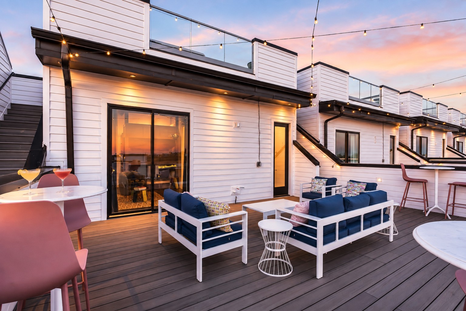 Rooftop deck with white lounge seating, pink dining chairs, and string lights at sunset.