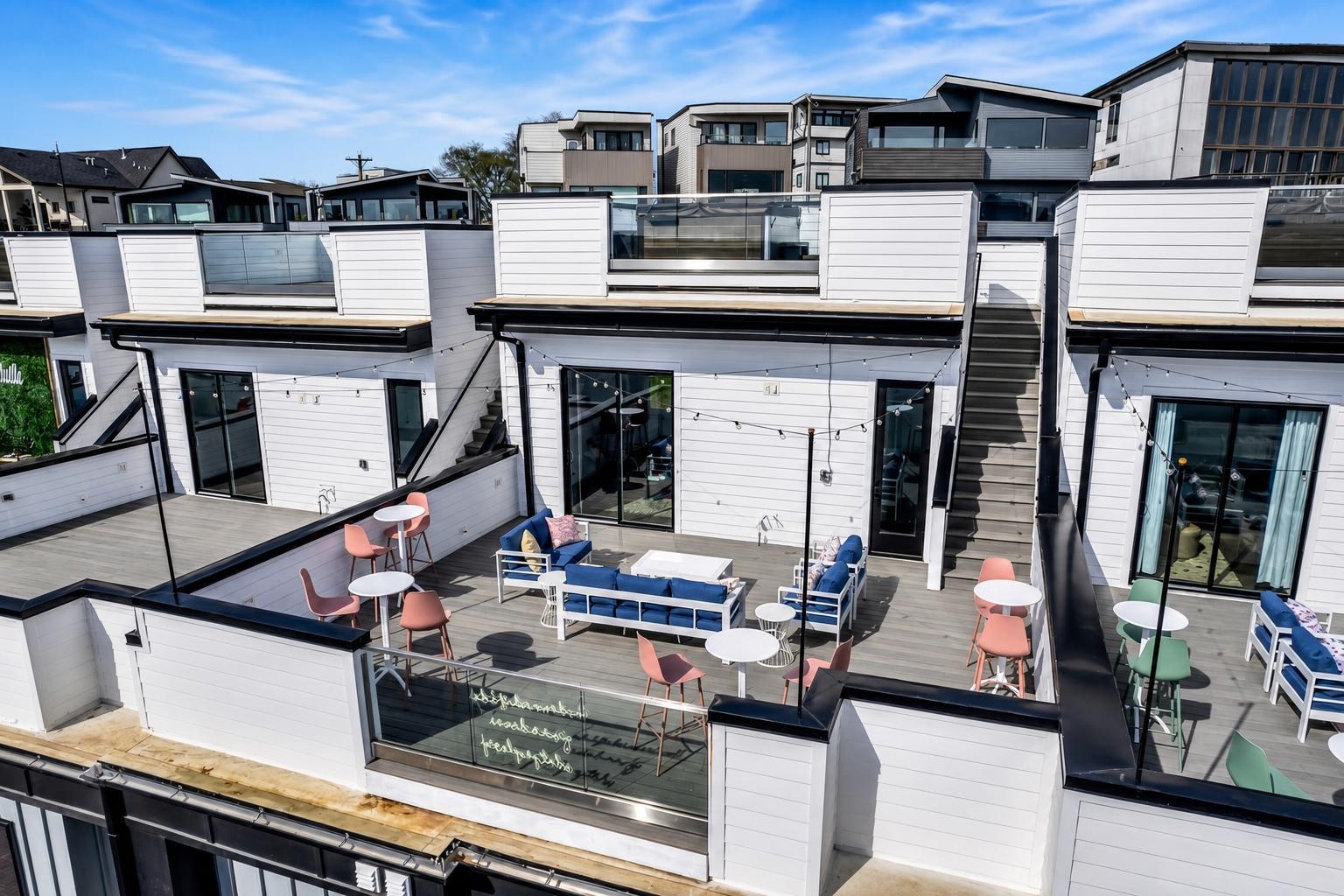 Furnished rooftop terrace with glass railings, blue lounge seating, dining tables, and stairs between modern townhomes.